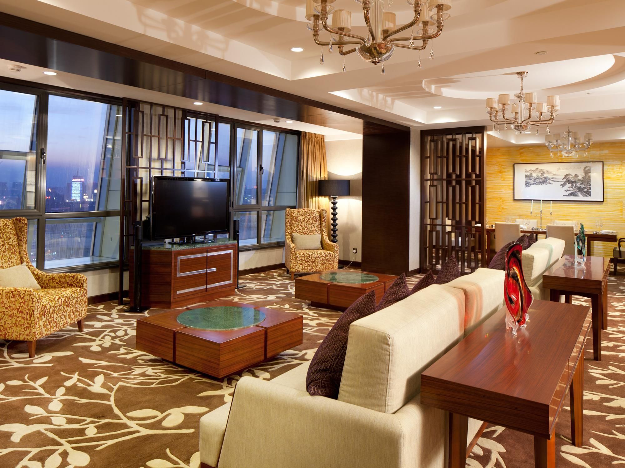 A modern hotel lobby with a large TV, comfortable seating, a dining area, and a view of the cityscape.