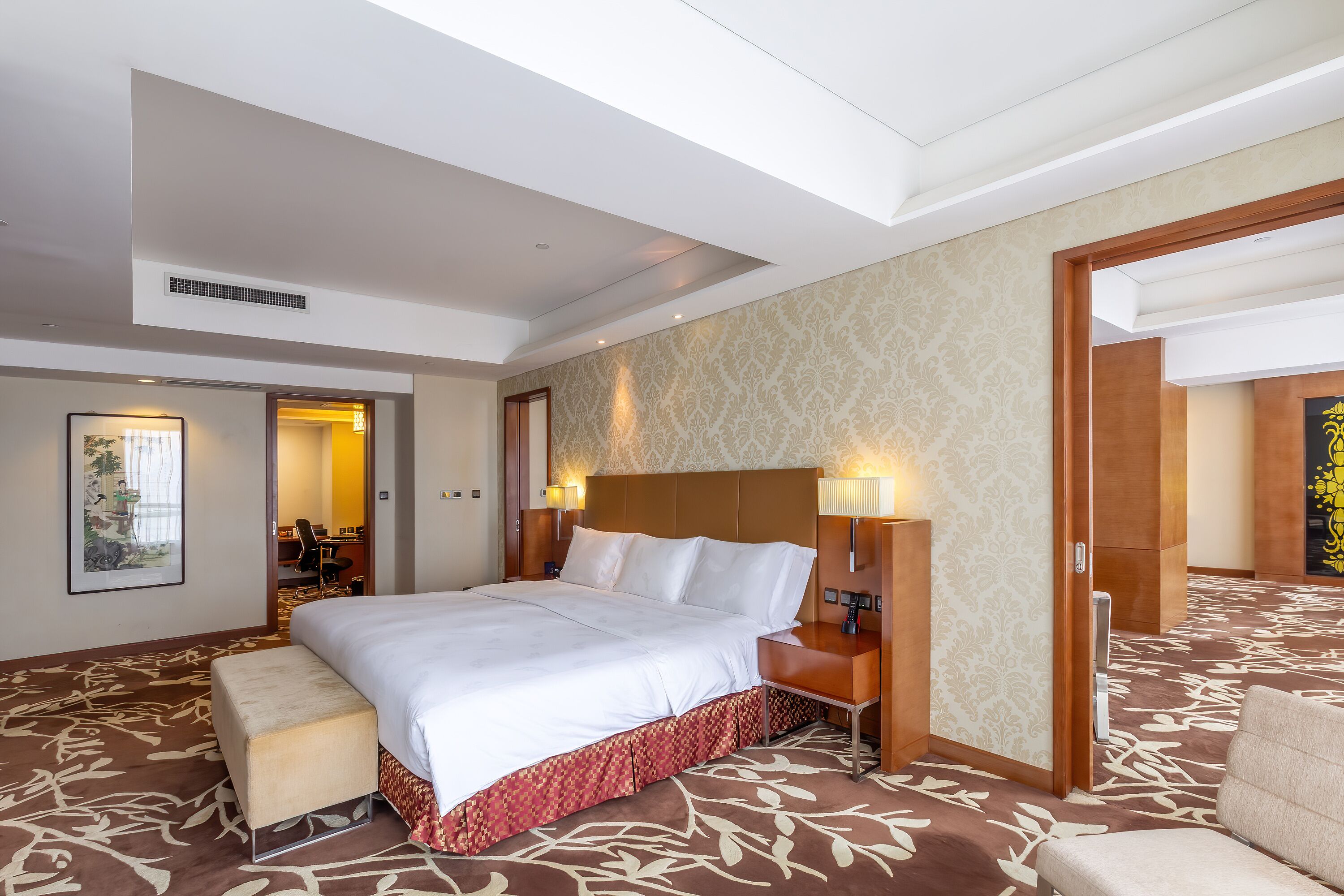 A hotel room with a large bed, a wooden headboard, a bench, and a carpeted floor.