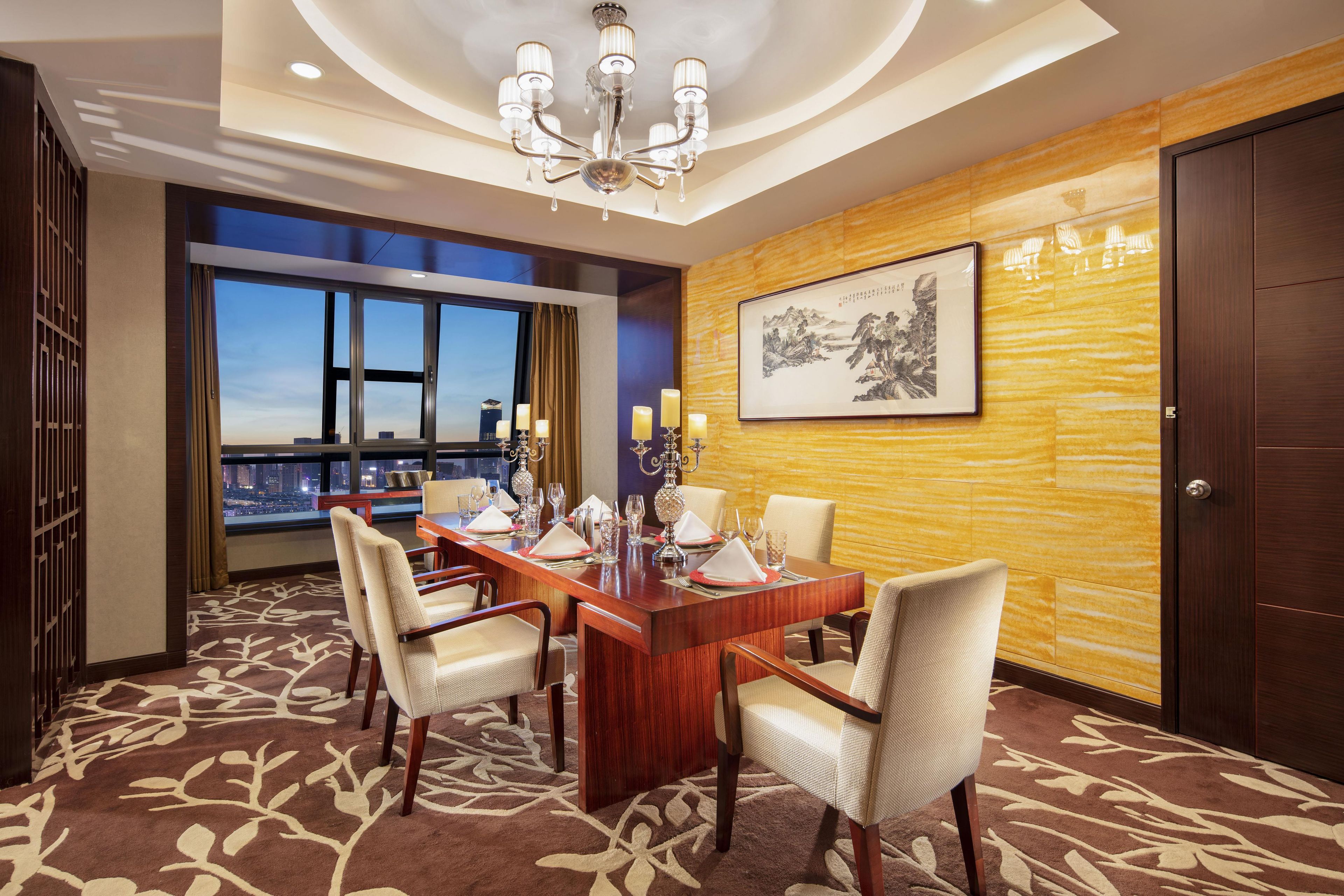 Een hotelconferentieruimte met een lange houten tafel, stoelen, een kroonluchter, uitzicht over de stad en een kunstwerk in een lijst aan de muur.