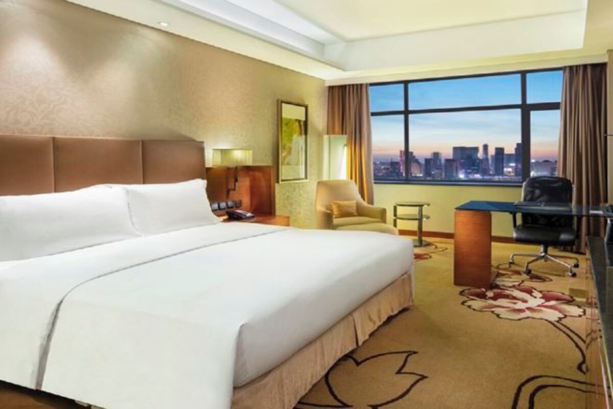 Habitación de hotel con una cama grande, un escritorio, una silla y vistas a un horizonte urbano a través de un ventanal.