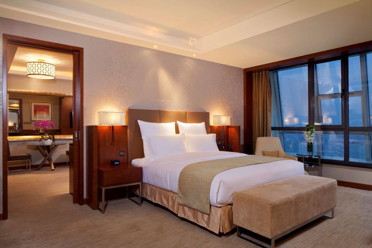 A hotel room with a large bed, bedside tables, a desk, and a view of the city through large windows.