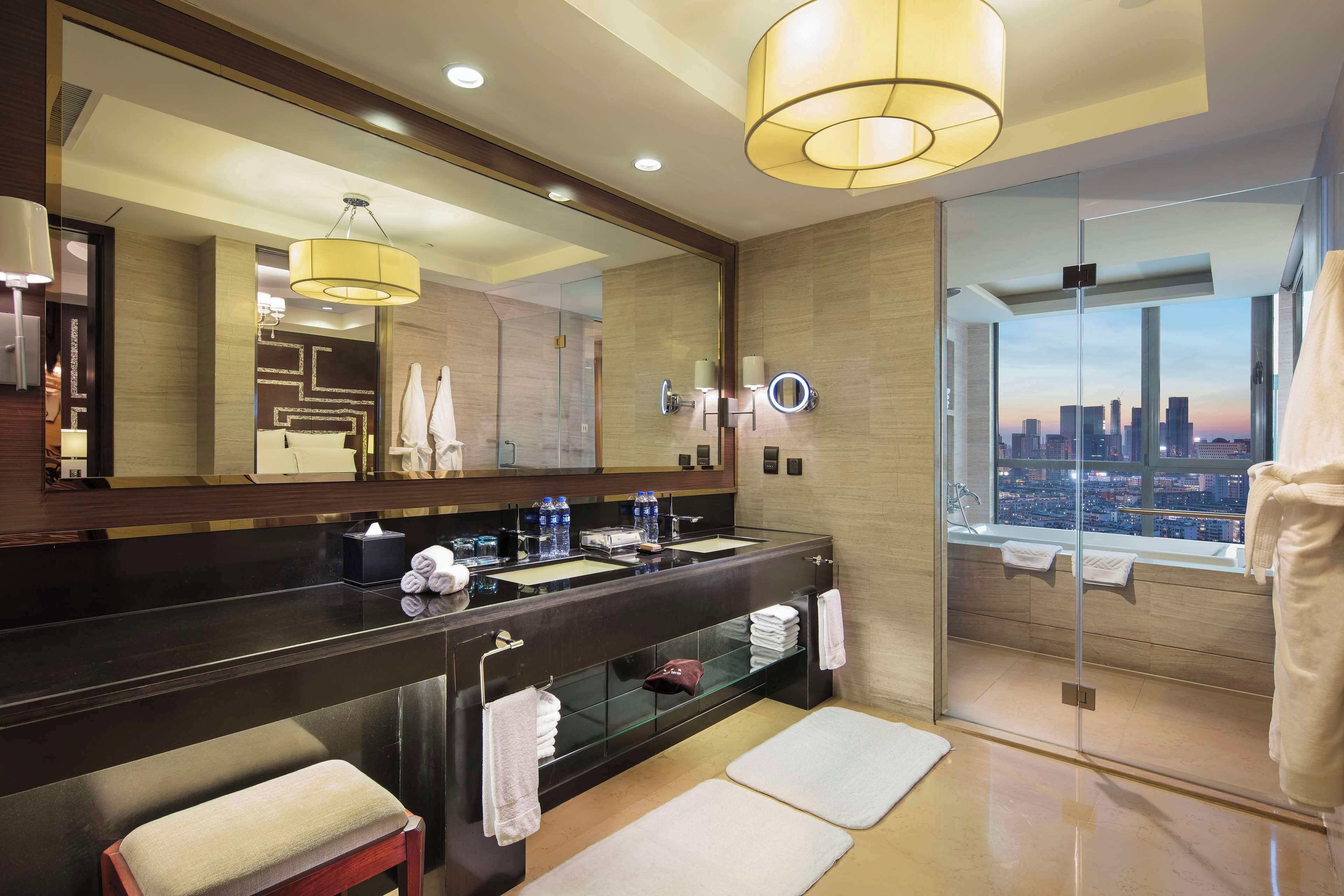 A modern bathroom with a large mirror, dual sinks, a glass shower enclosure, and a city view.