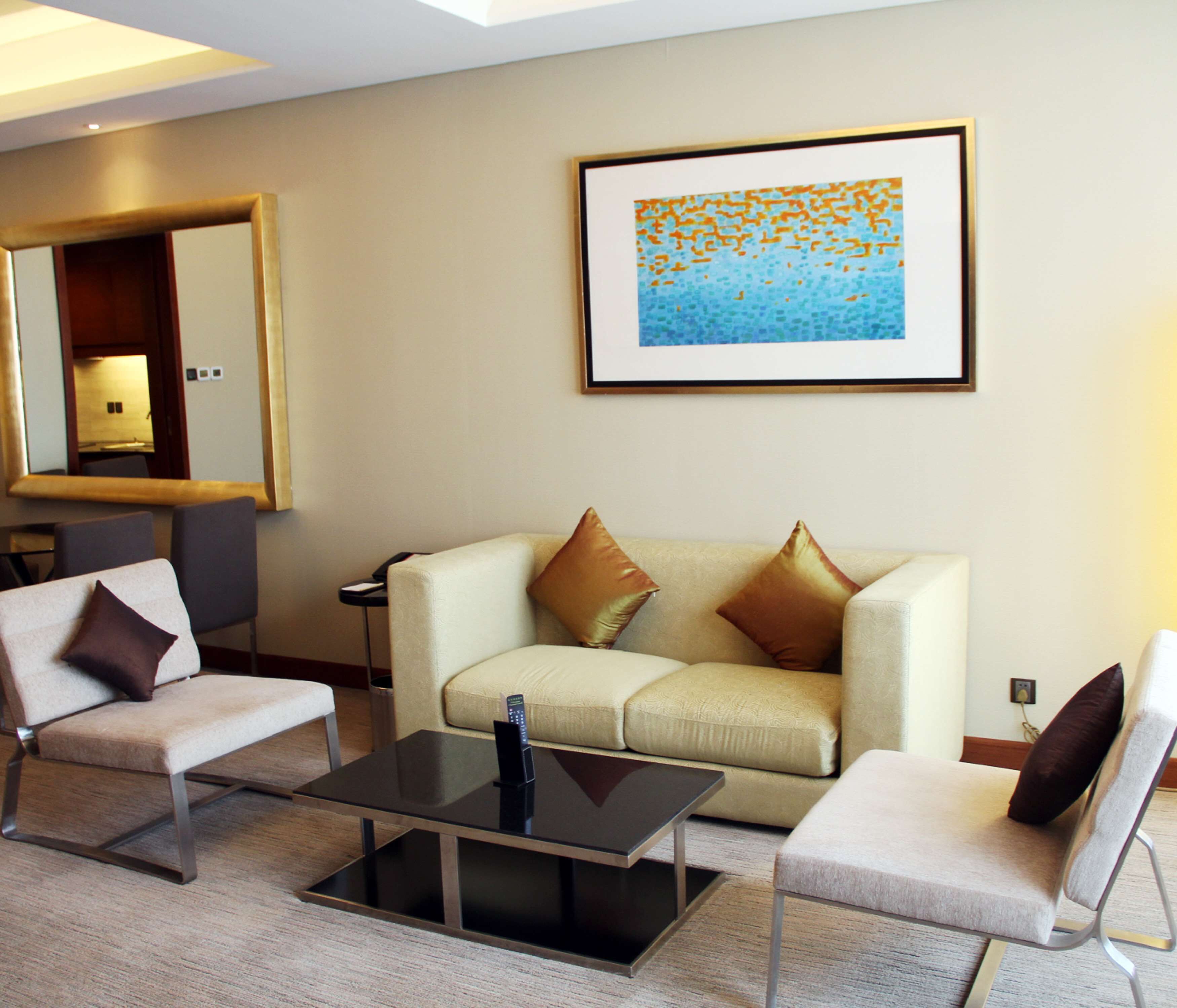 A modern hotel room with a beige sofa, two armchairs, a glass coffee table, and a framed abstract painting on the wall.
