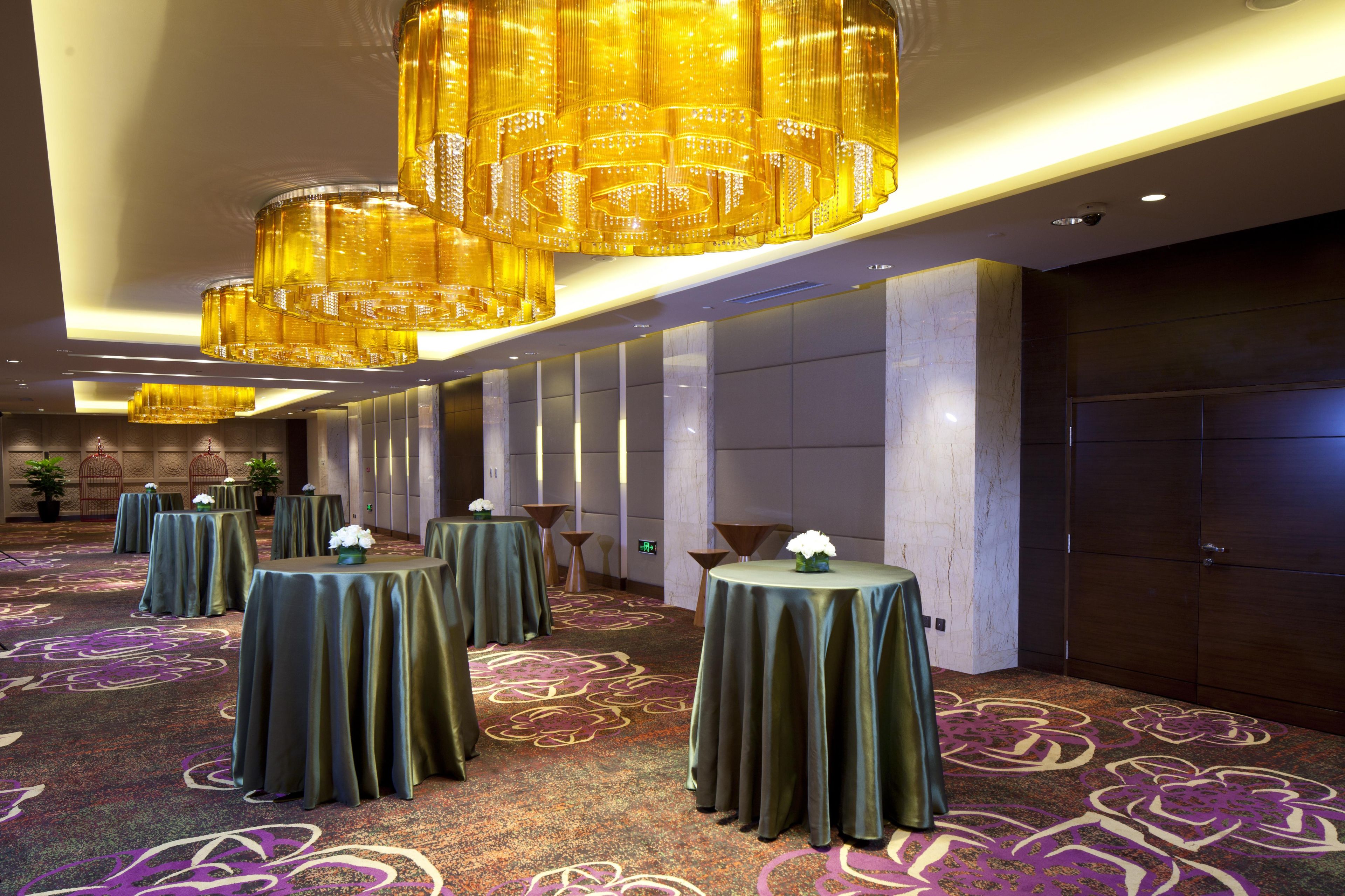 A hotel lobby with multiple round tables covered with green tablecloths, large golden chandeliers, and a patterned carpet.