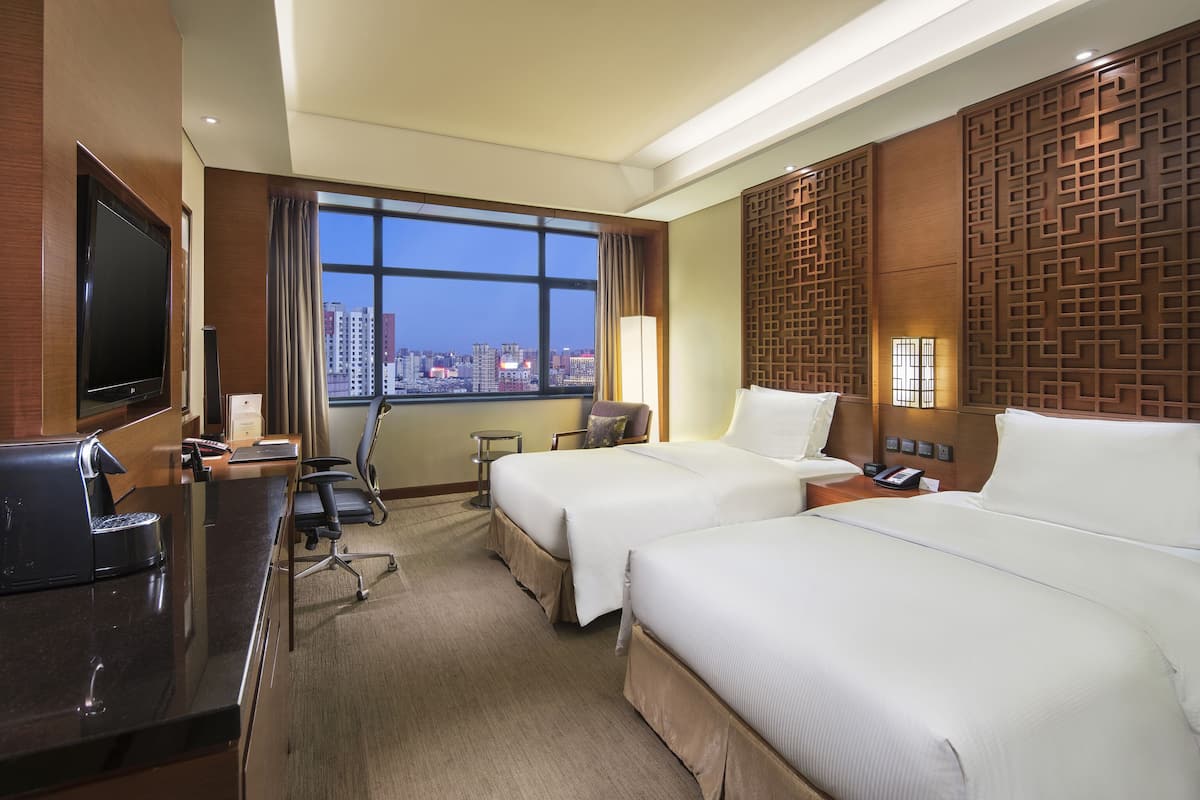 A hotel room with two beds, a desk with a coffee maker, a TV, and a large window with a city view.