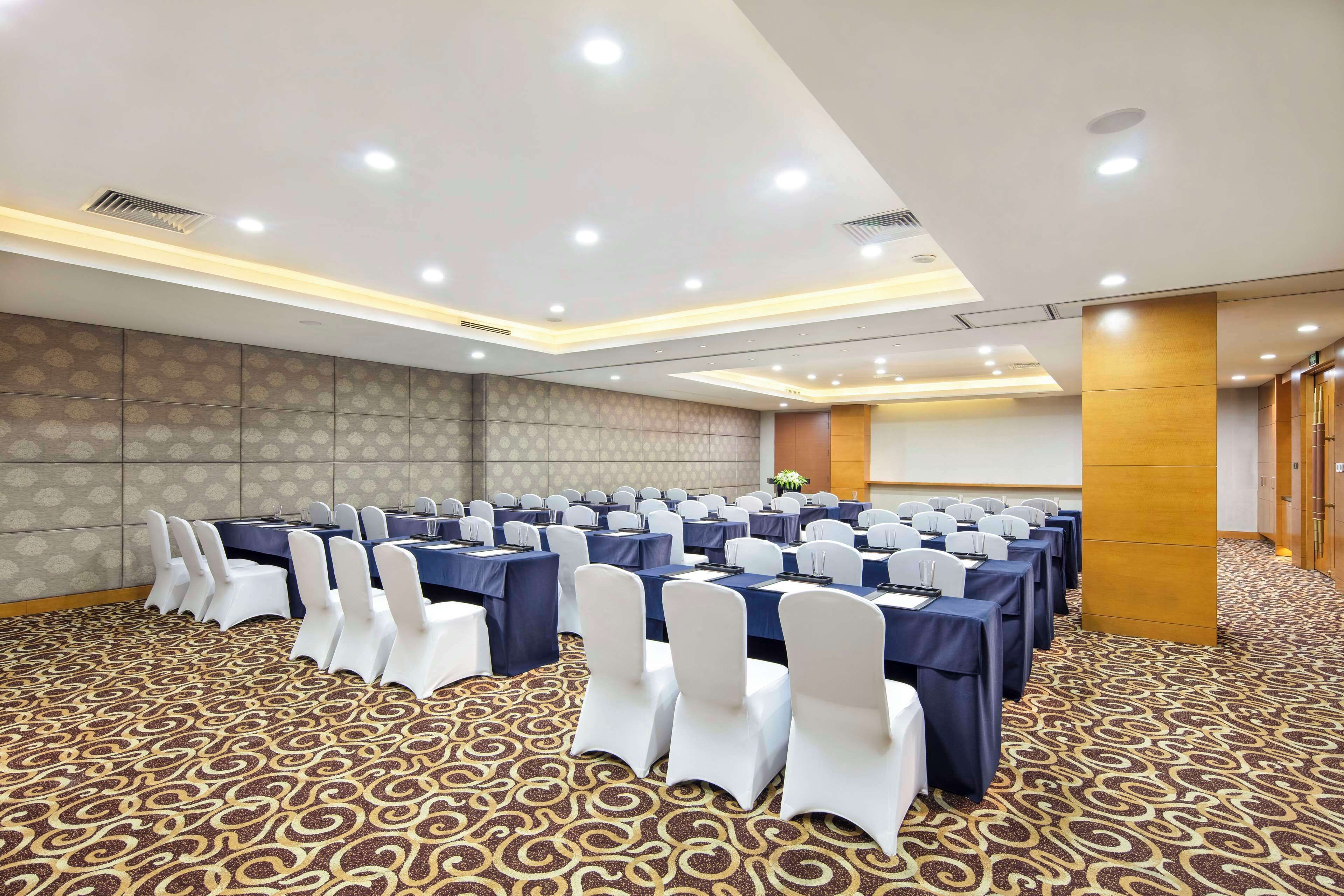 A conference room with rows of tables and chairs, a patterned carpet, and recessed ceiling lighting.