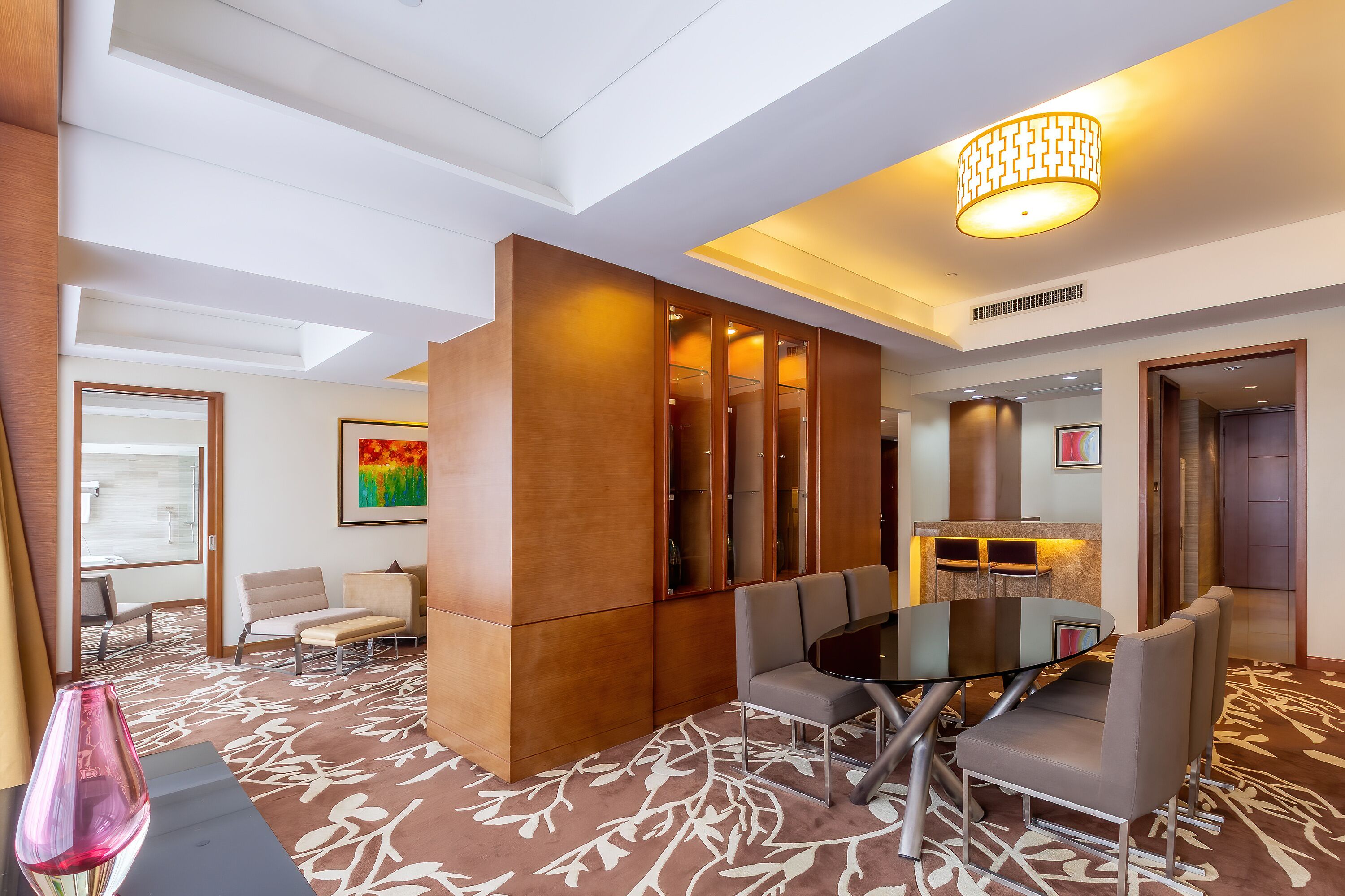 A modern hotel lobby with a glass table, cushioned chairs, a patterned carpet, and a large wooden partition.