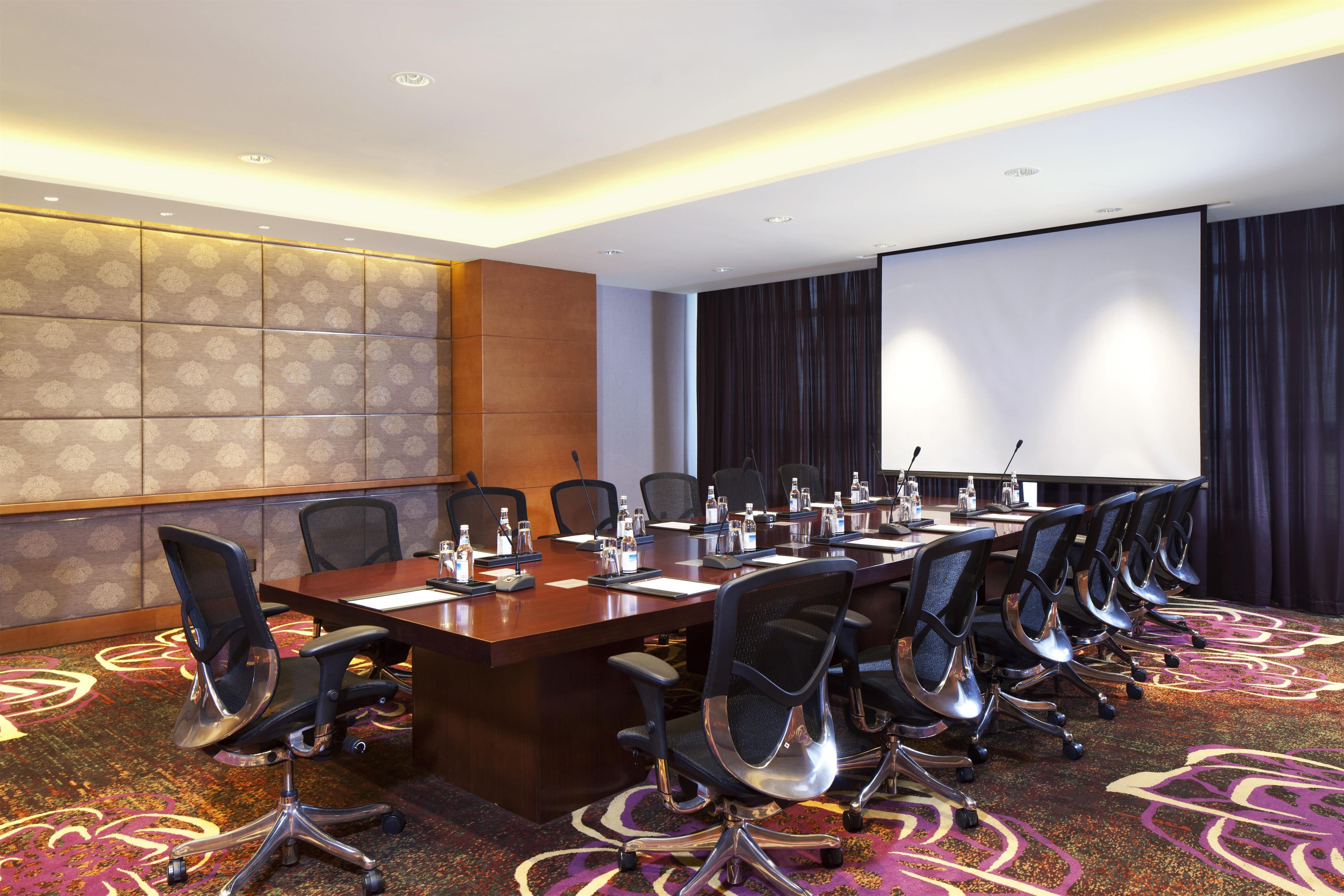 A conference room with a long table, multiple chairs, a projector screen, and a carpeted floor.