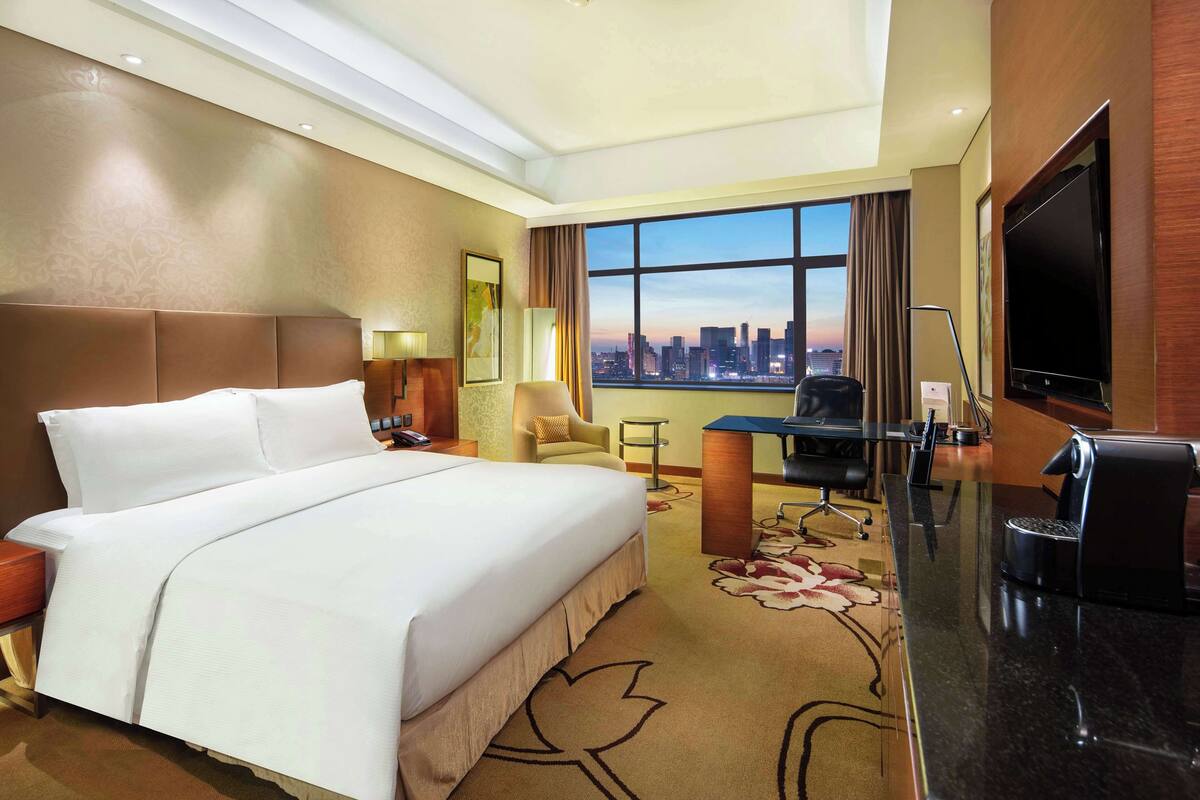 A hotel room with a large bed, a desk with a chair, a TV, and a city view through the window.