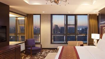 A hotel room with a large bed, a TV, a chair, a desk, and a view of the cityscape through the windows.