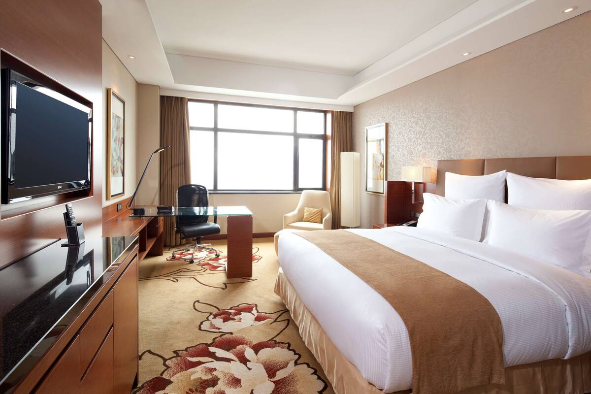A hotel room with a large bed, a desk with a computer, a TV, and a window with curtains.