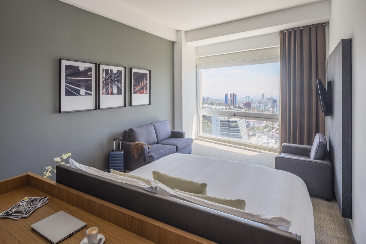 A modern hotel room with a large bed, a sofa, a desk with a laptop and coffee cup, a TV mounted on the wall, and framed pictures on the wall.