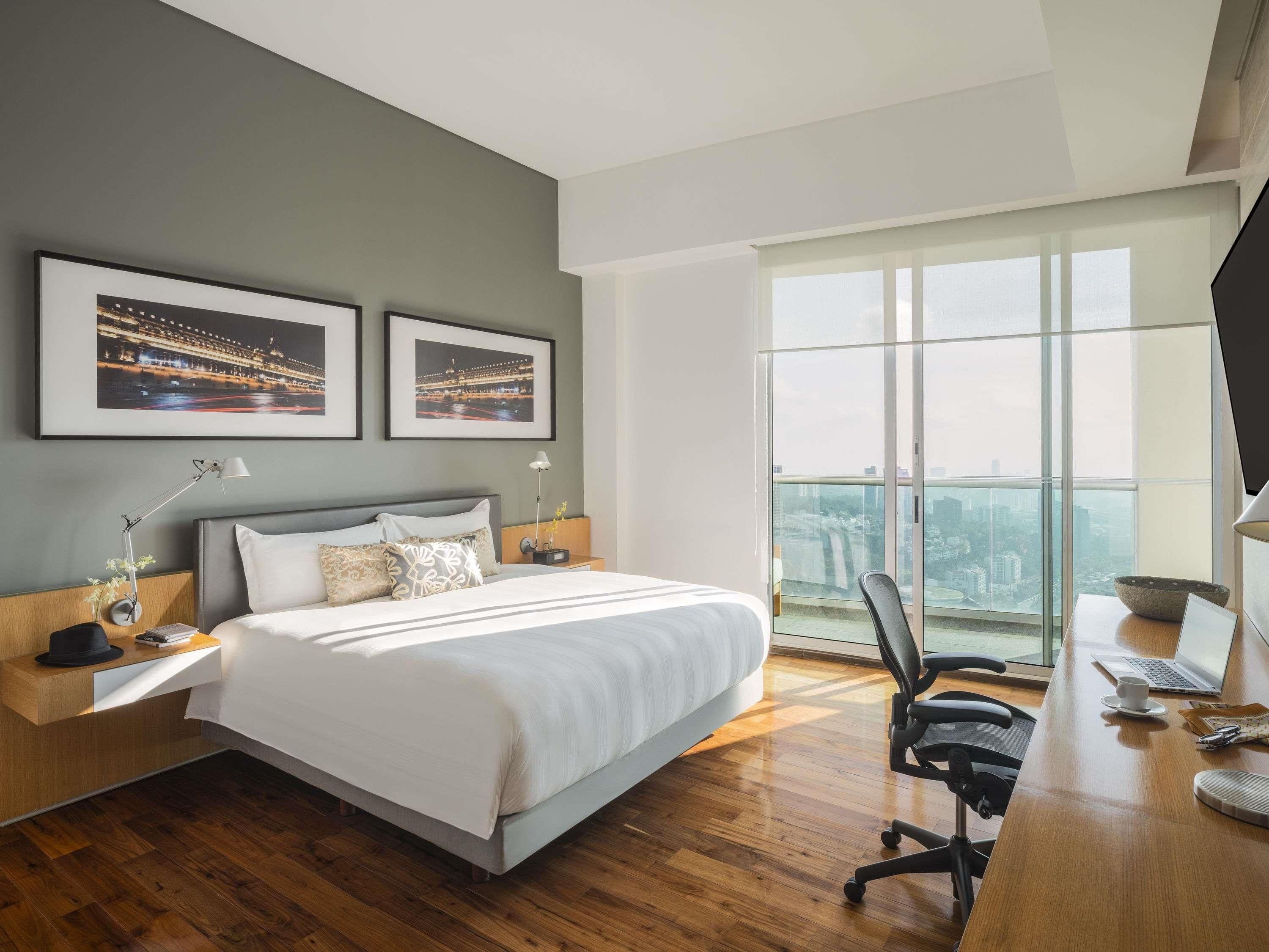 A modern hotel room with a large bed, a desk with a laptop, and a city view through floor-to-ceiling windows.