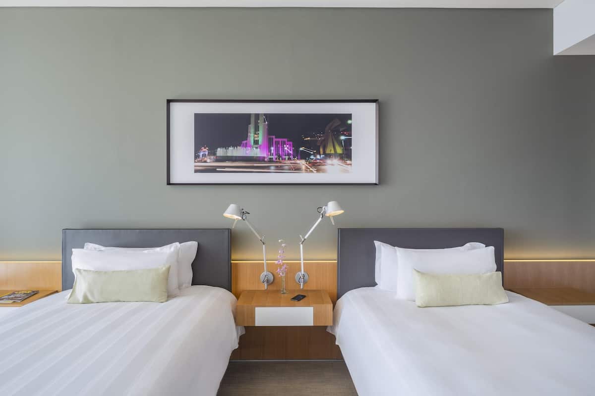 A hotel room with two beds, a nightstand with lamps, and a framed cityscape picture on the wall.