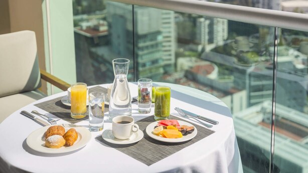 A table with breakfast items, a glass of orange juice, water, and a green smoothie, with a cityscape view through a glass railing. - The Paragon Hotel Mexico City Santa Fe By Accor (Mexico City)