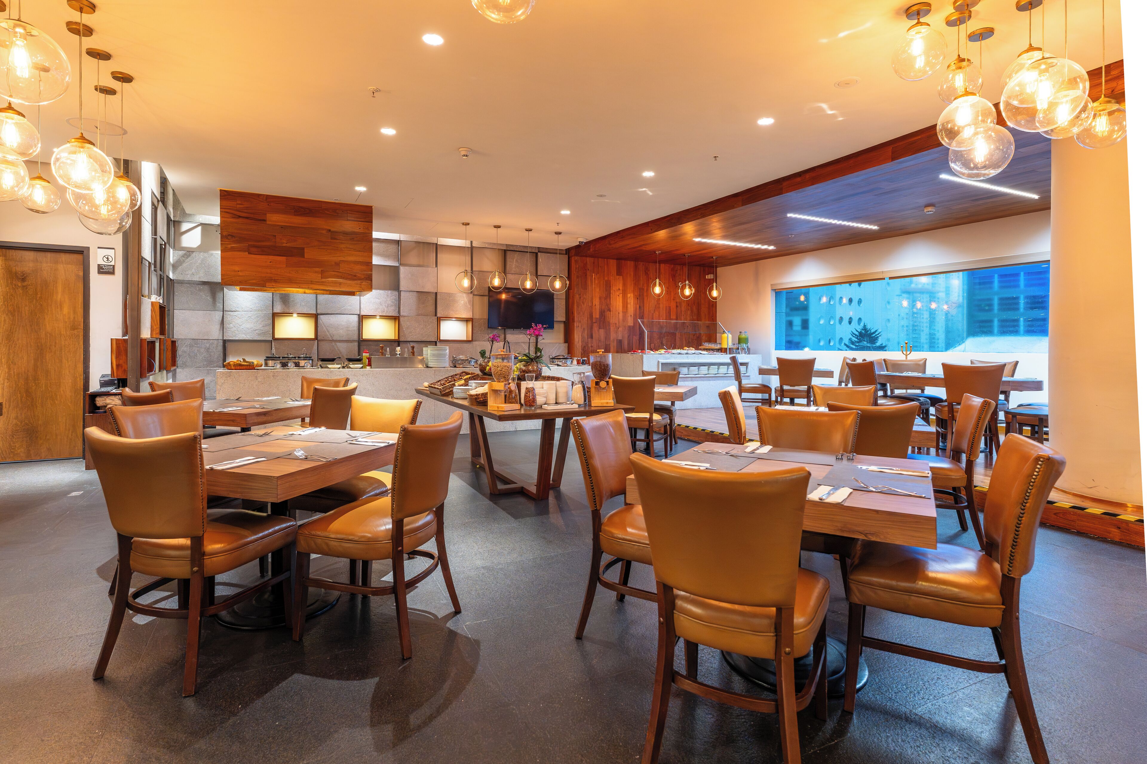 A modern restaurant with wooden tables and chairs, a kitchen area, and large windows offering a view of the cityscape.