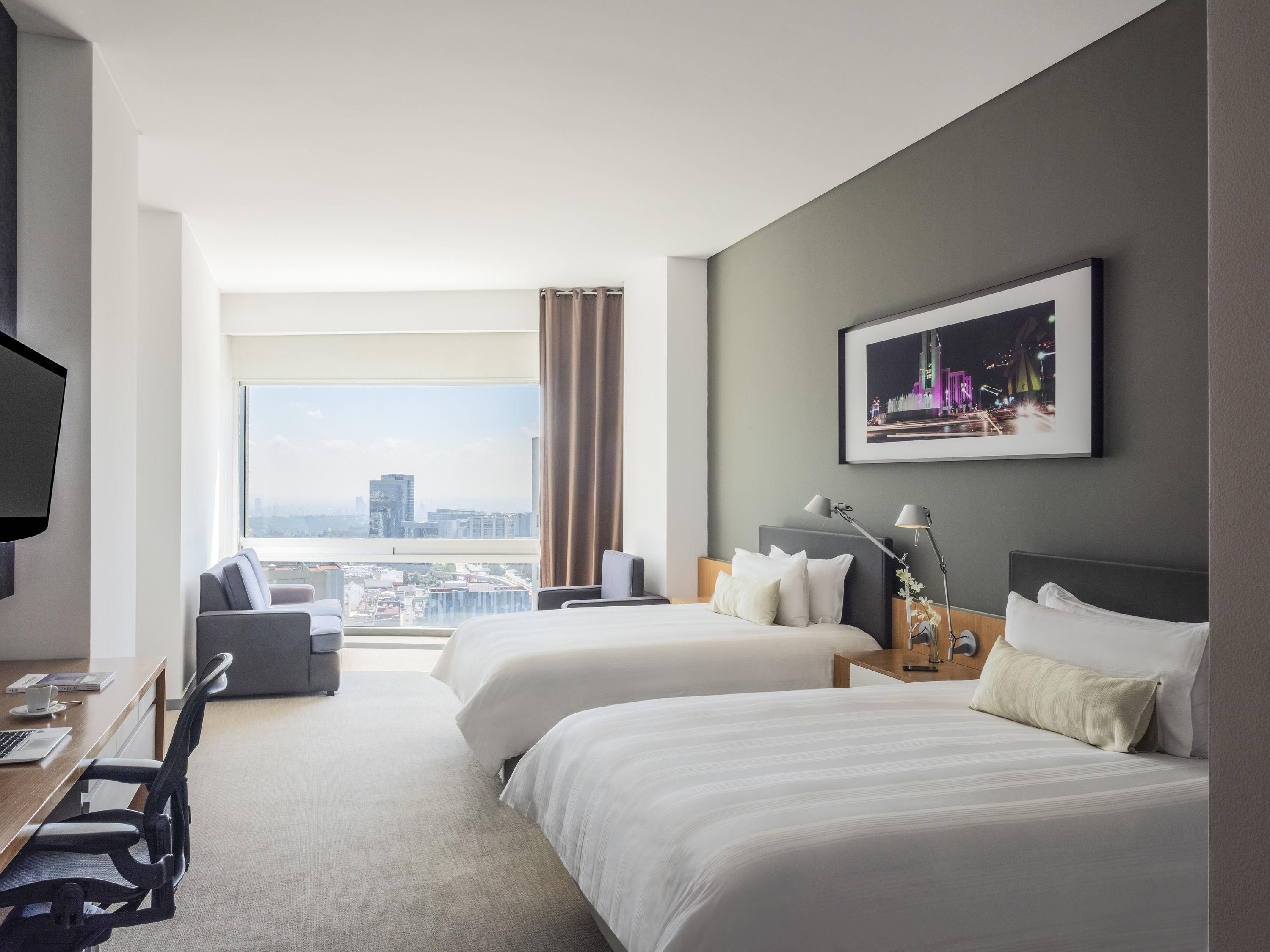 A hotel room with two beds, a desk, a chair, a TV, and a large window offering a city view.