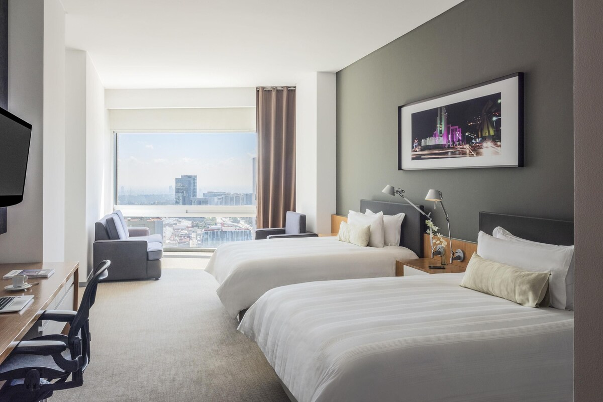 A hotel room with two beds, a desk, a chair, a TV, and a large window offering a city view.