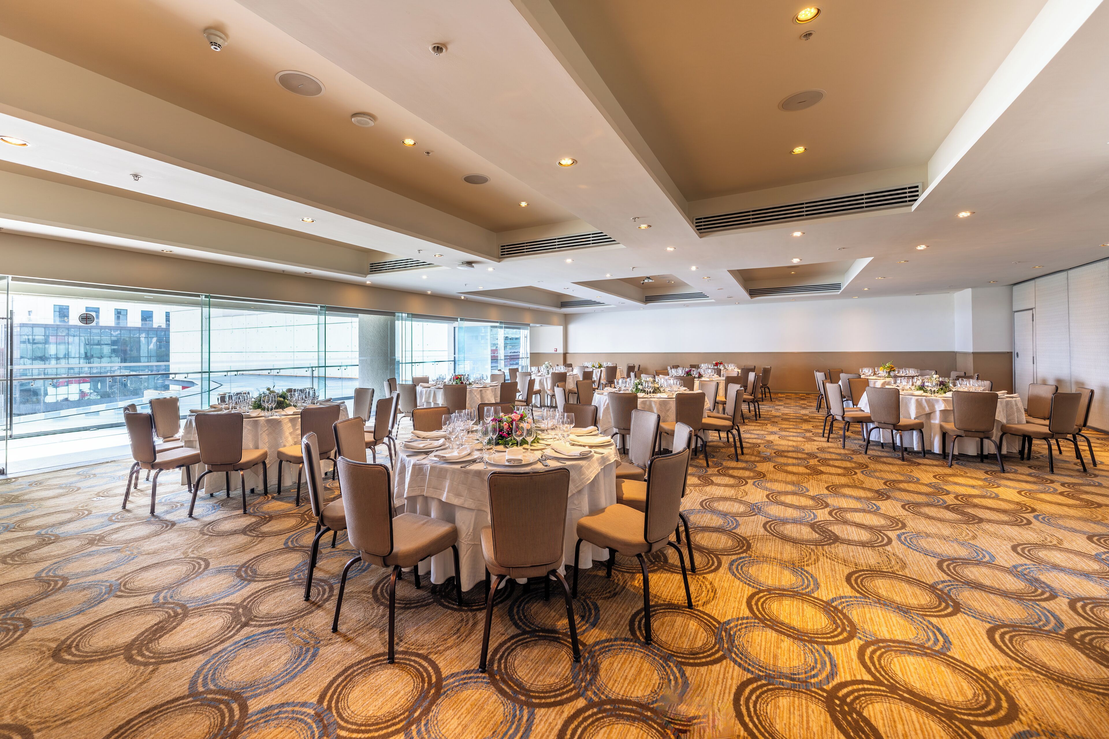 Une grande salle de conférence dotée de plusieurs tables rondes disposées pour un repas, avec des chaises et des tables, et recouverte d’un tapis à motifs.
