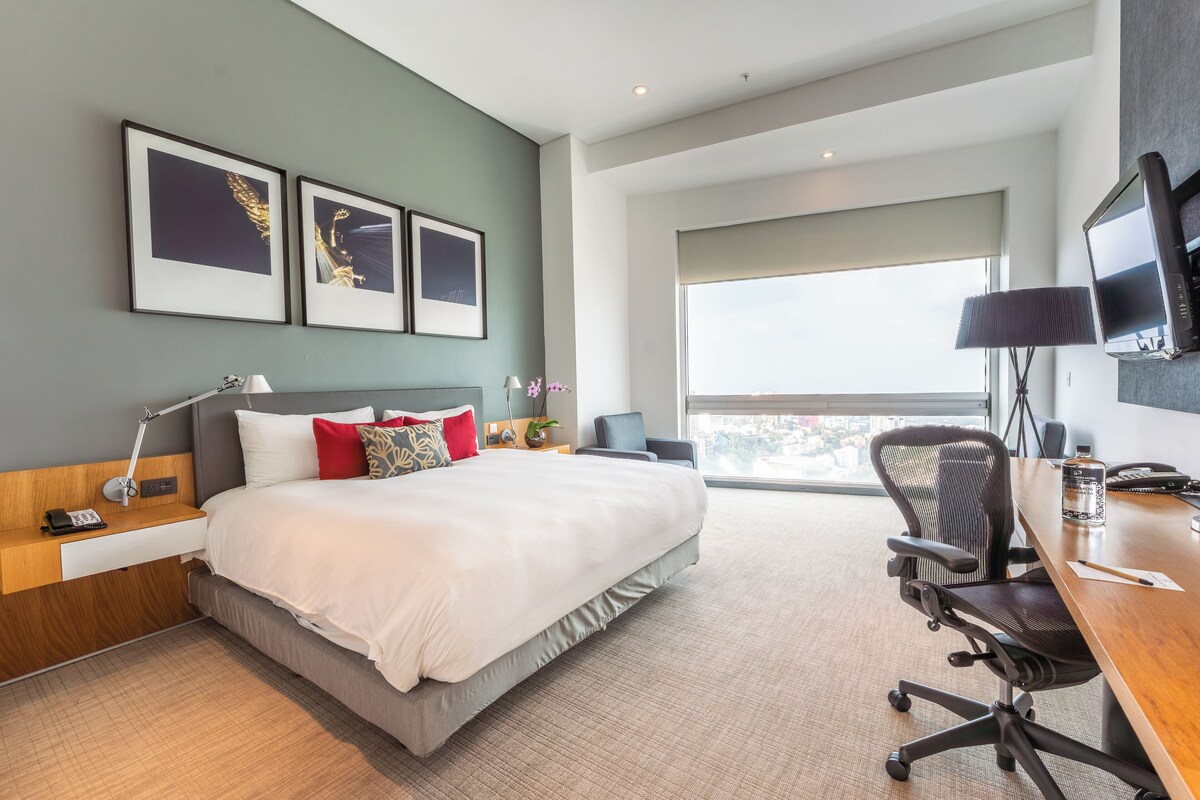 A modern hotel room with a large bed, a desk with a chair, a TV mounted on the wall, and a large window offering a city view.