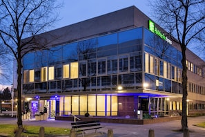 Un hôtel Holiday Inn Express doté d’une façade moderne en verre, d’un espace extérieur aménagé pour s’asseoir et d’une entrée bien éclairée.