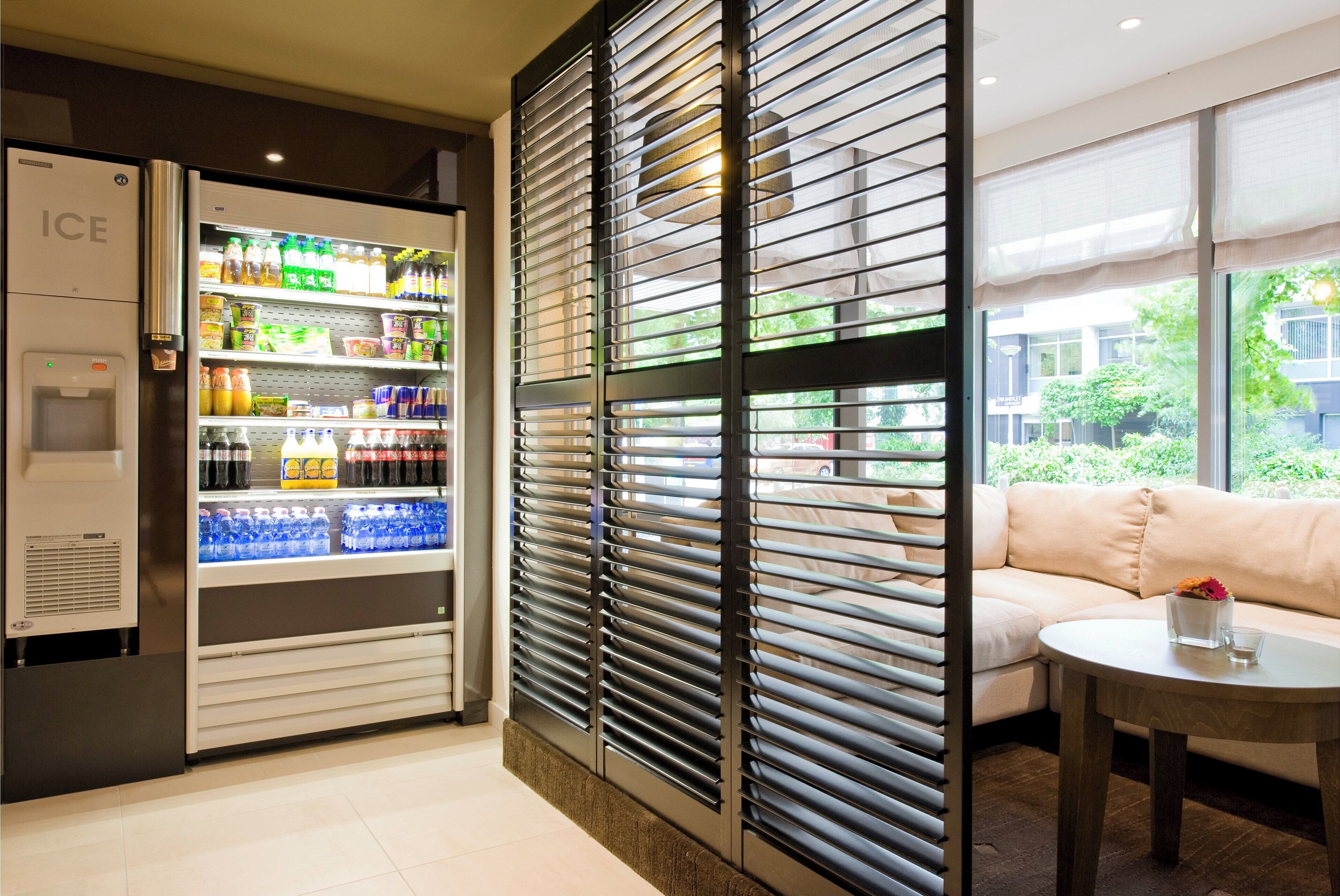 A hotel room with a minibar, a sofa, a table, and a window with a view of greenery.