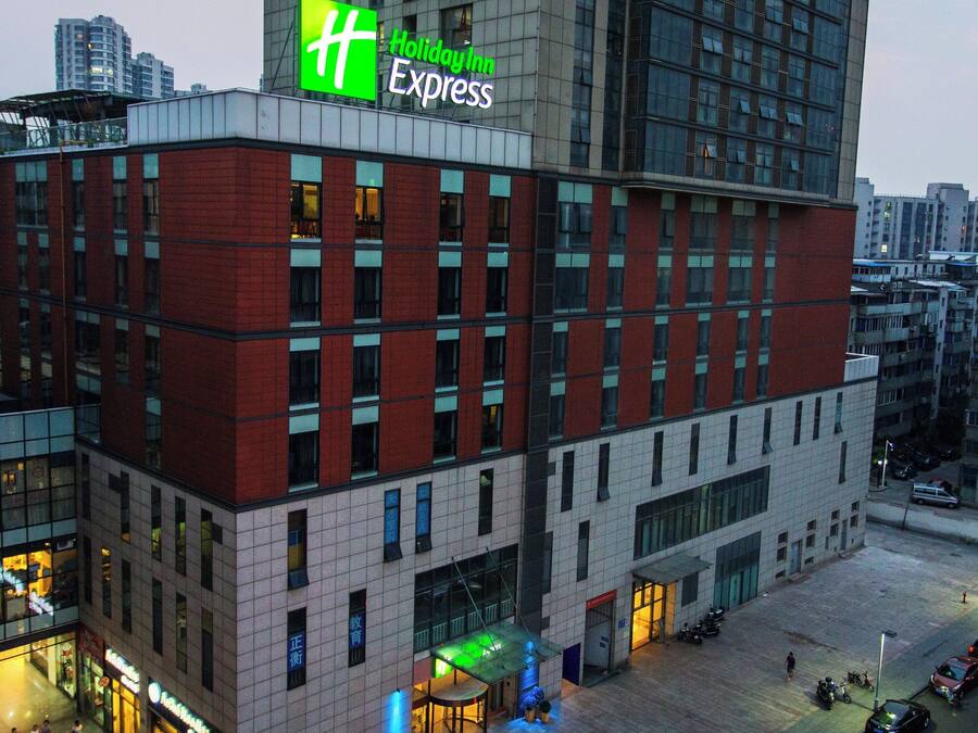 A Holiday Inn Express hotel with a modern architectural design, featuring a prominent green and white sign, multiple windows, and a parking area visible below.