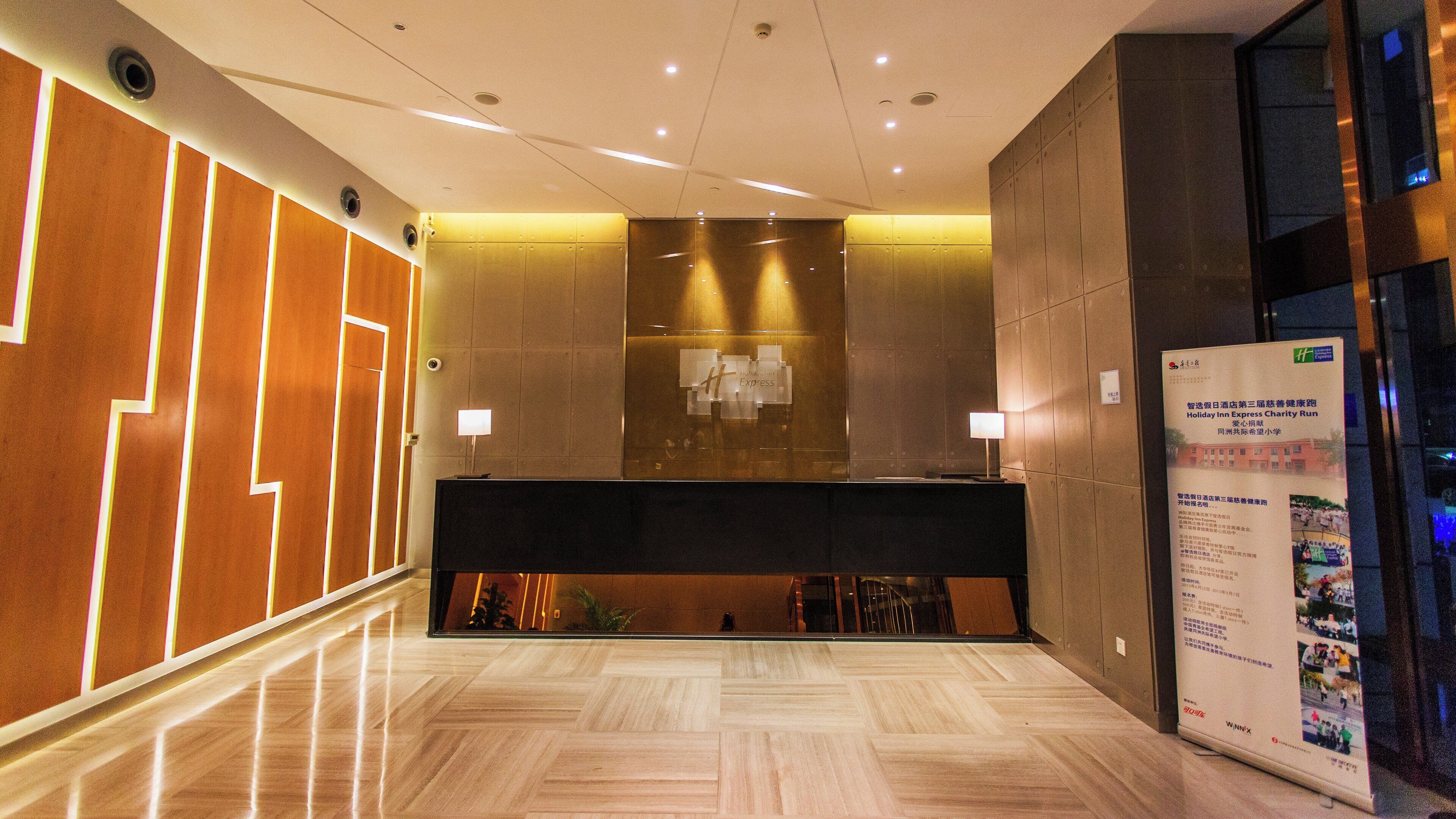 A hotel lobby with a reception desk, a wall-mounted information board, and a wooden floor.