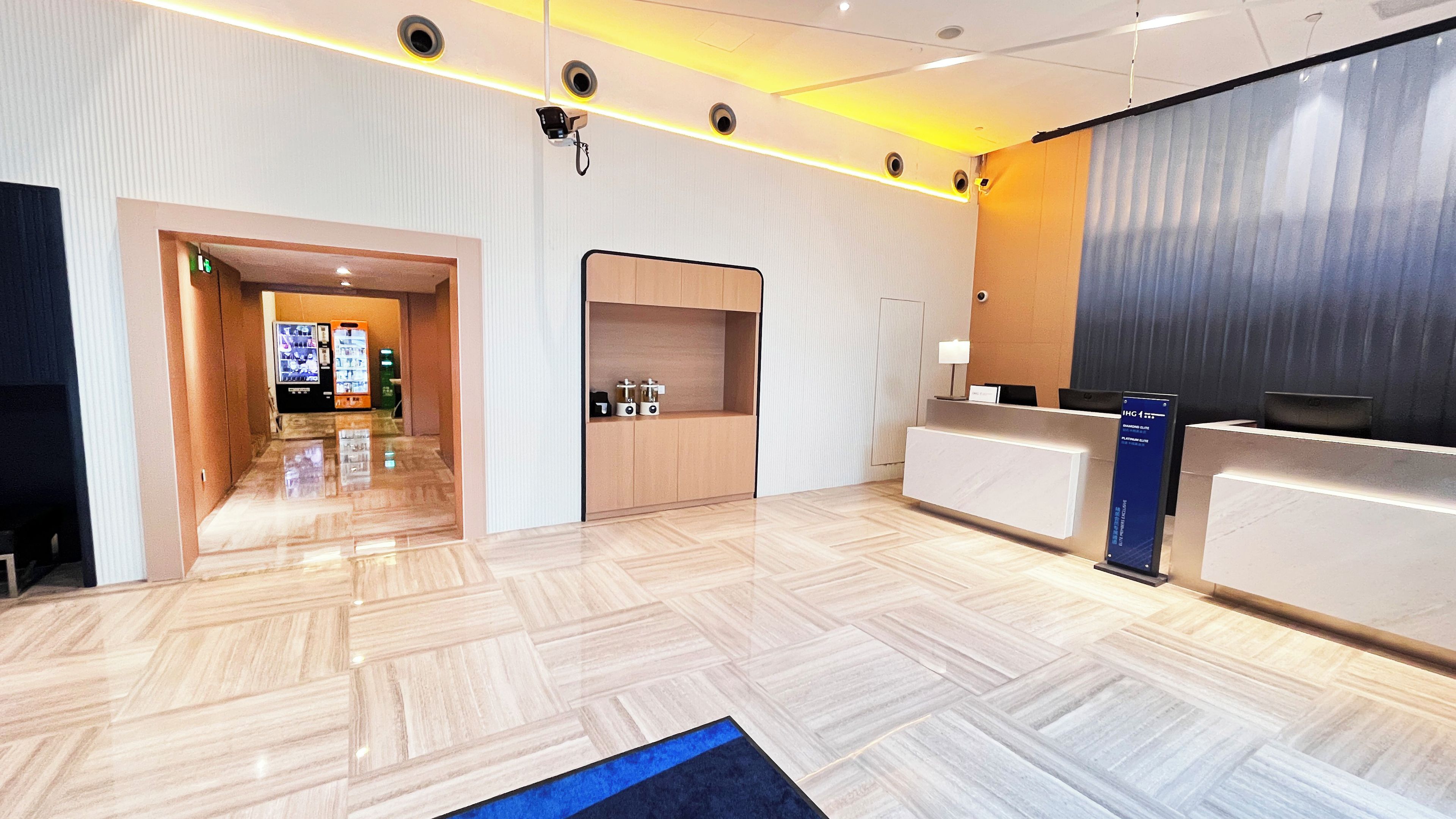 Zona de recepción de hotel con diseño moderno, que incluye un mostrador de recepción, un panel informativo digital y un pasillo que conduce a otras habitaciones.