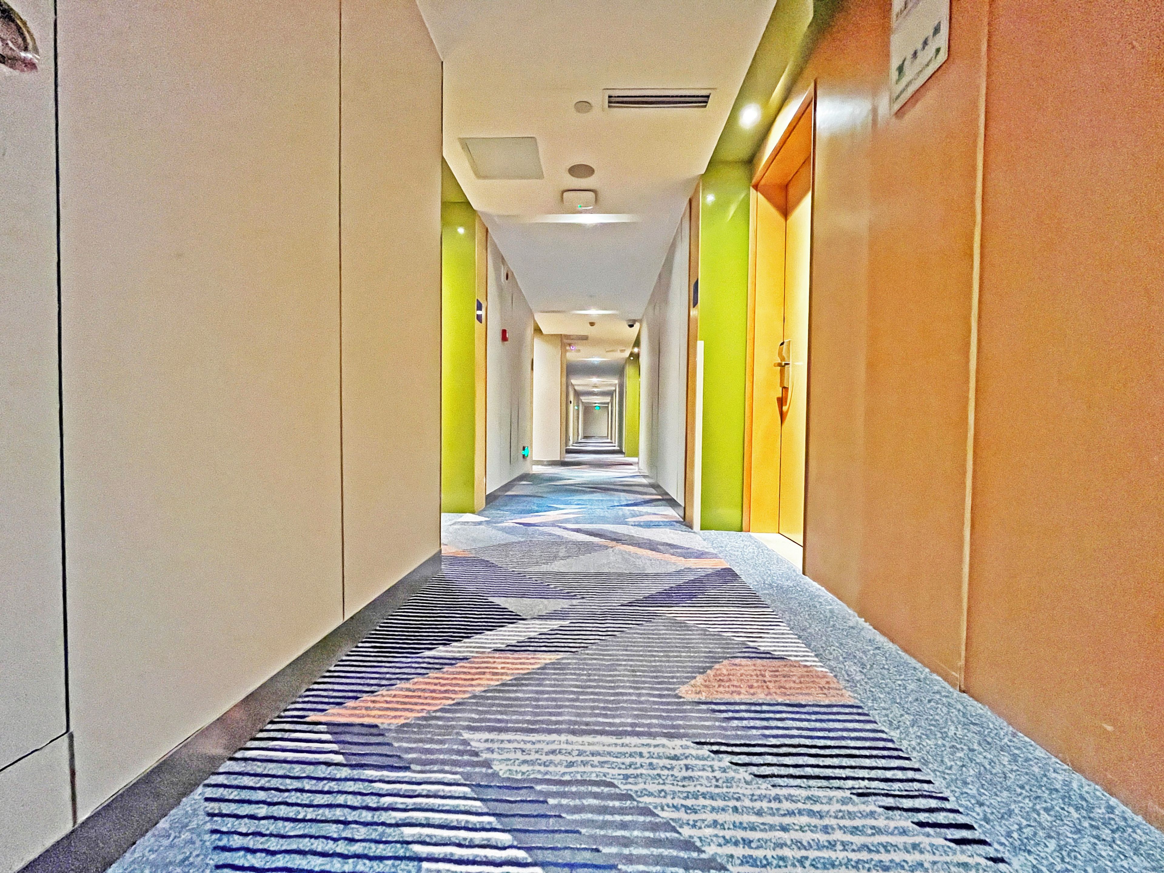 A hotel corridor with a patterned carpet, doors on the right, and a sign on the left.