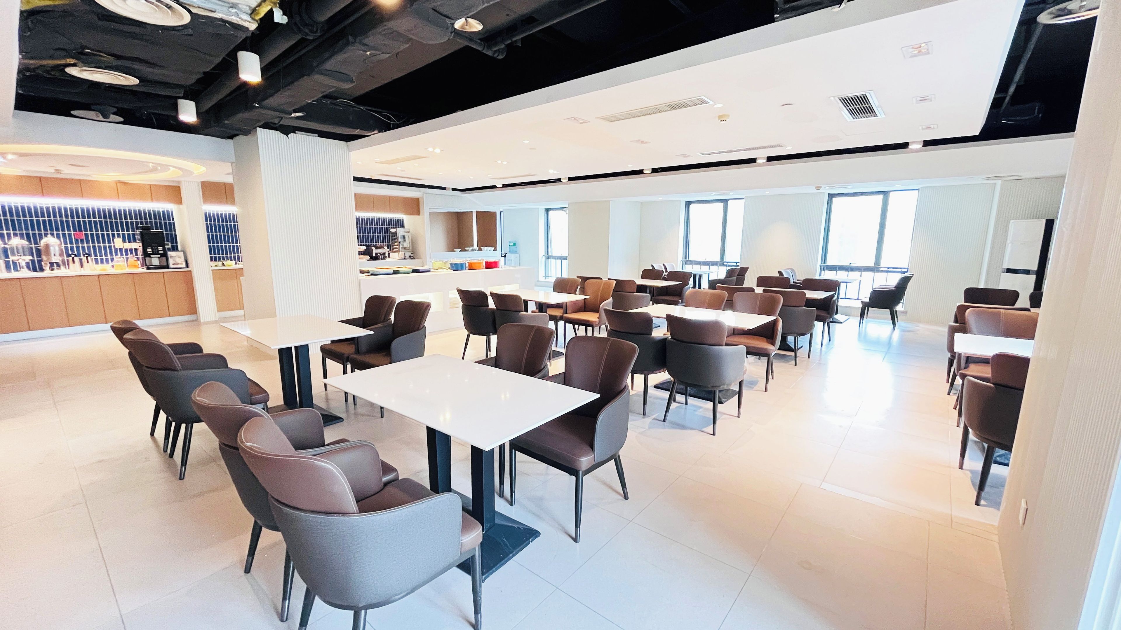 A modern dining area with multiple tables and chairs, a kitchen in the background, and large windows allowing natural light.