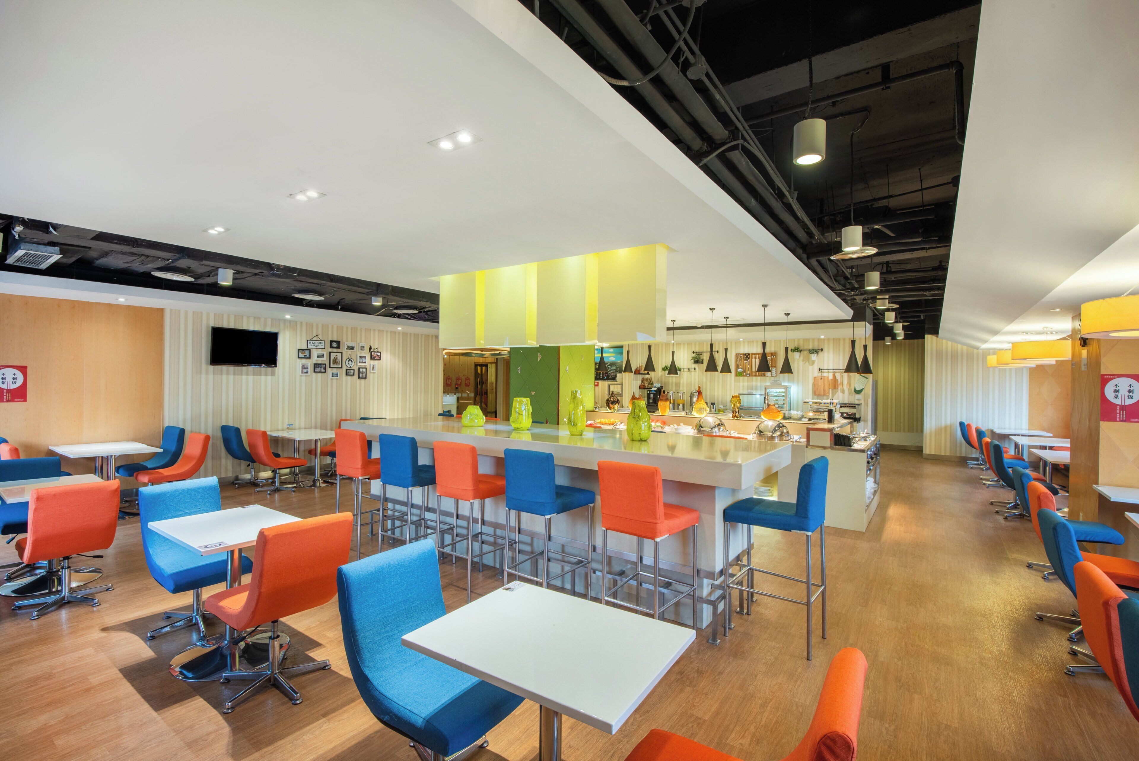 A modern cafeteria with high tables and colorful chairs, a central bar area, and a flat-screen TV mounted on the wall.