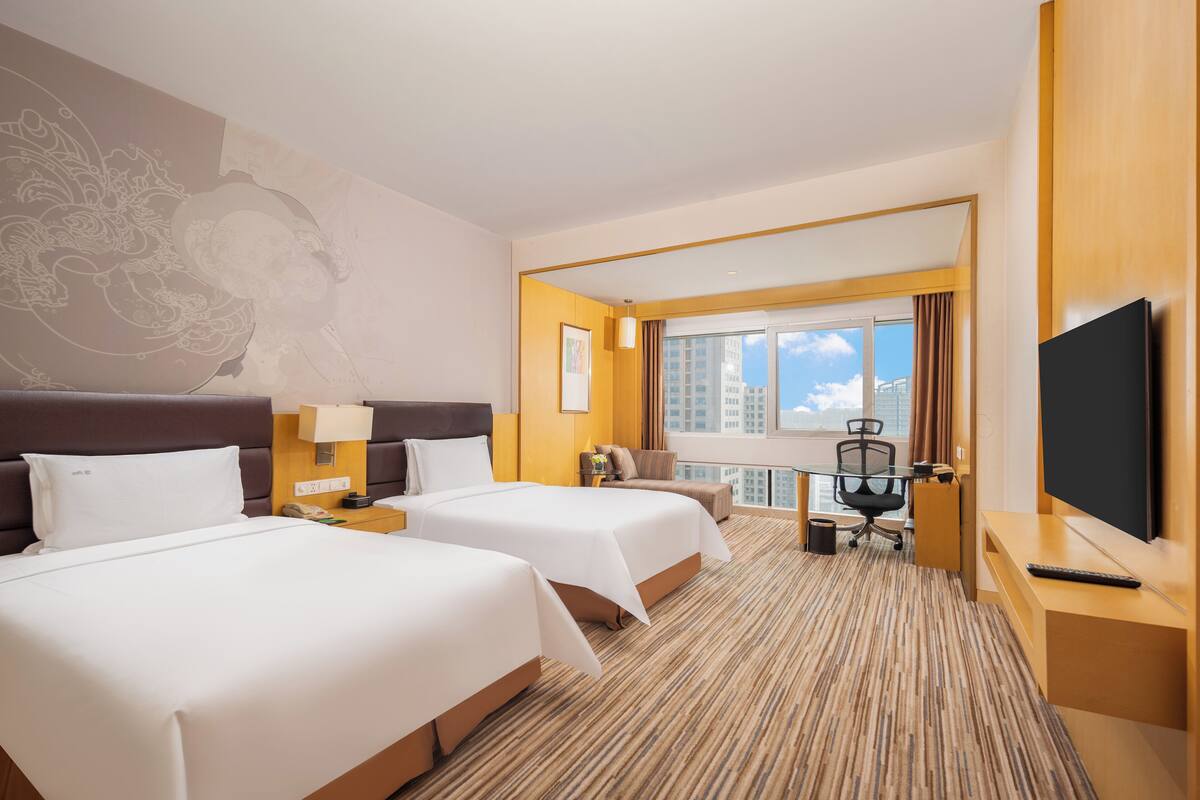 Ein Hotelzimmer mit zwei Betten, einem Schreibtisch, einem Fernseher und einem großen Fenster mit Blick auf die Stadt.