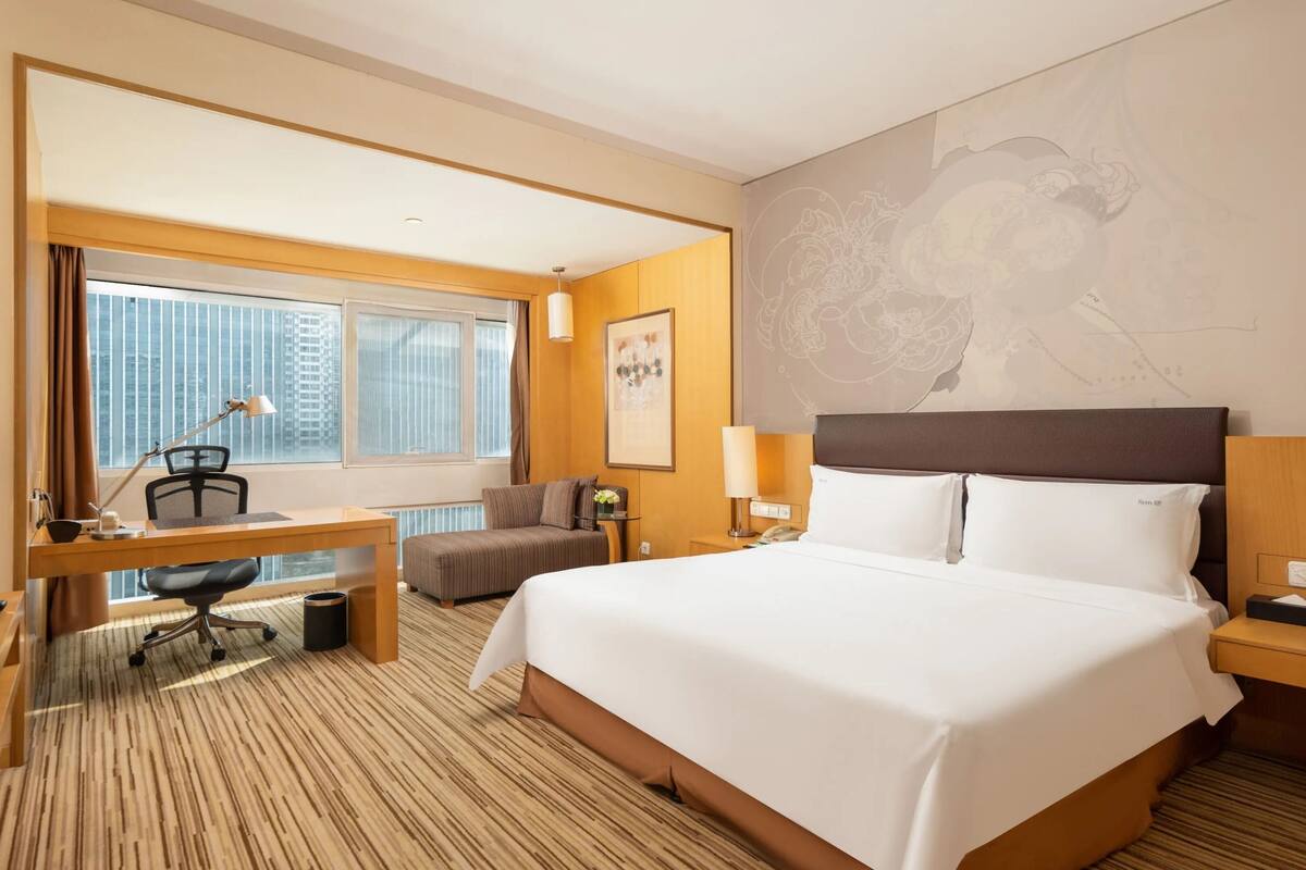 Ein Hotelzimmer mit einem großen Bett, einem Schreibtisch mit Stuhl, einem Fernseher und einem Fenster mit Vorhängen.