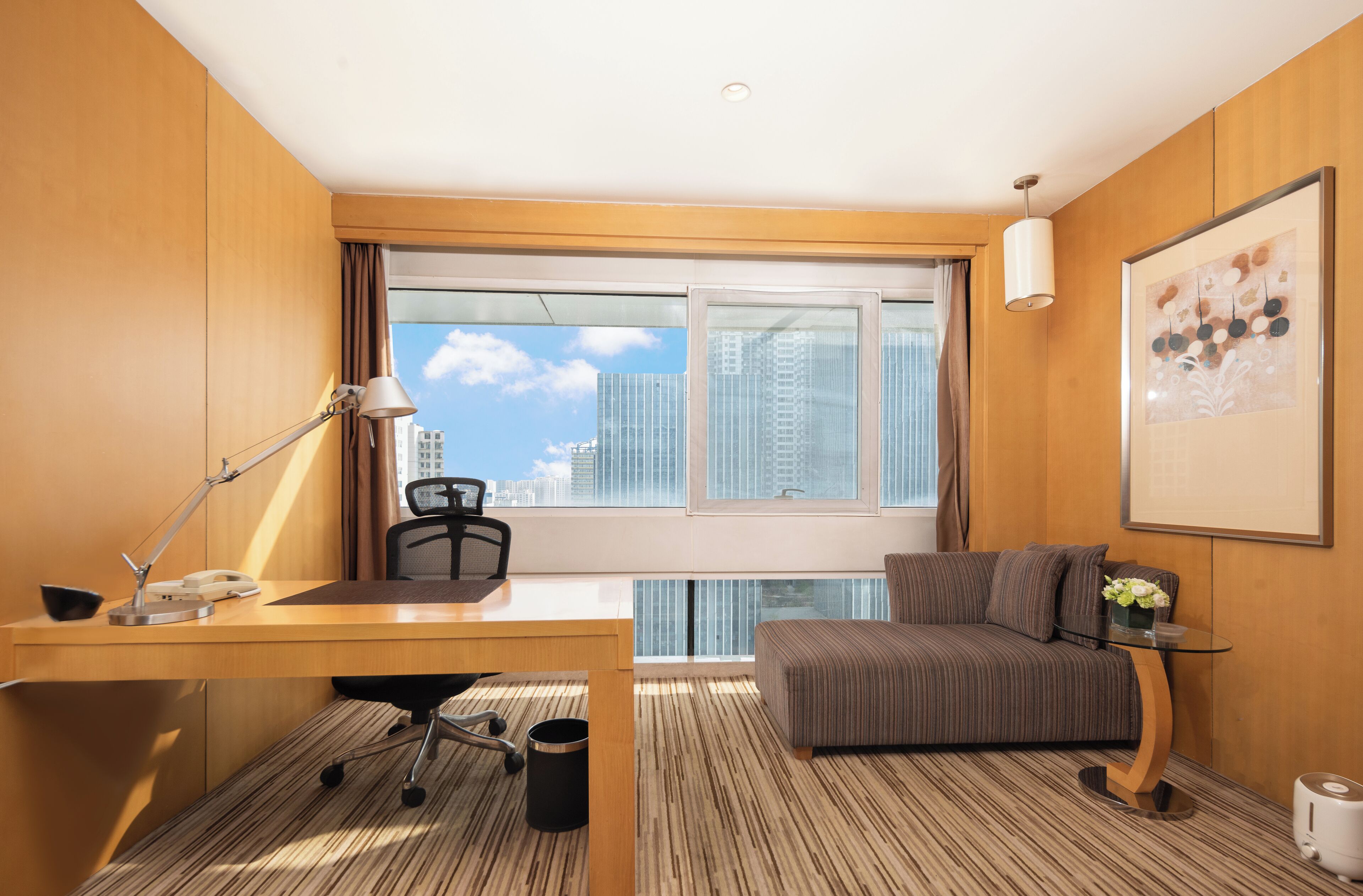 A hotel room with a wooden desk, a chair, a sofa, a small table with a vase, and a large window offering a city view.