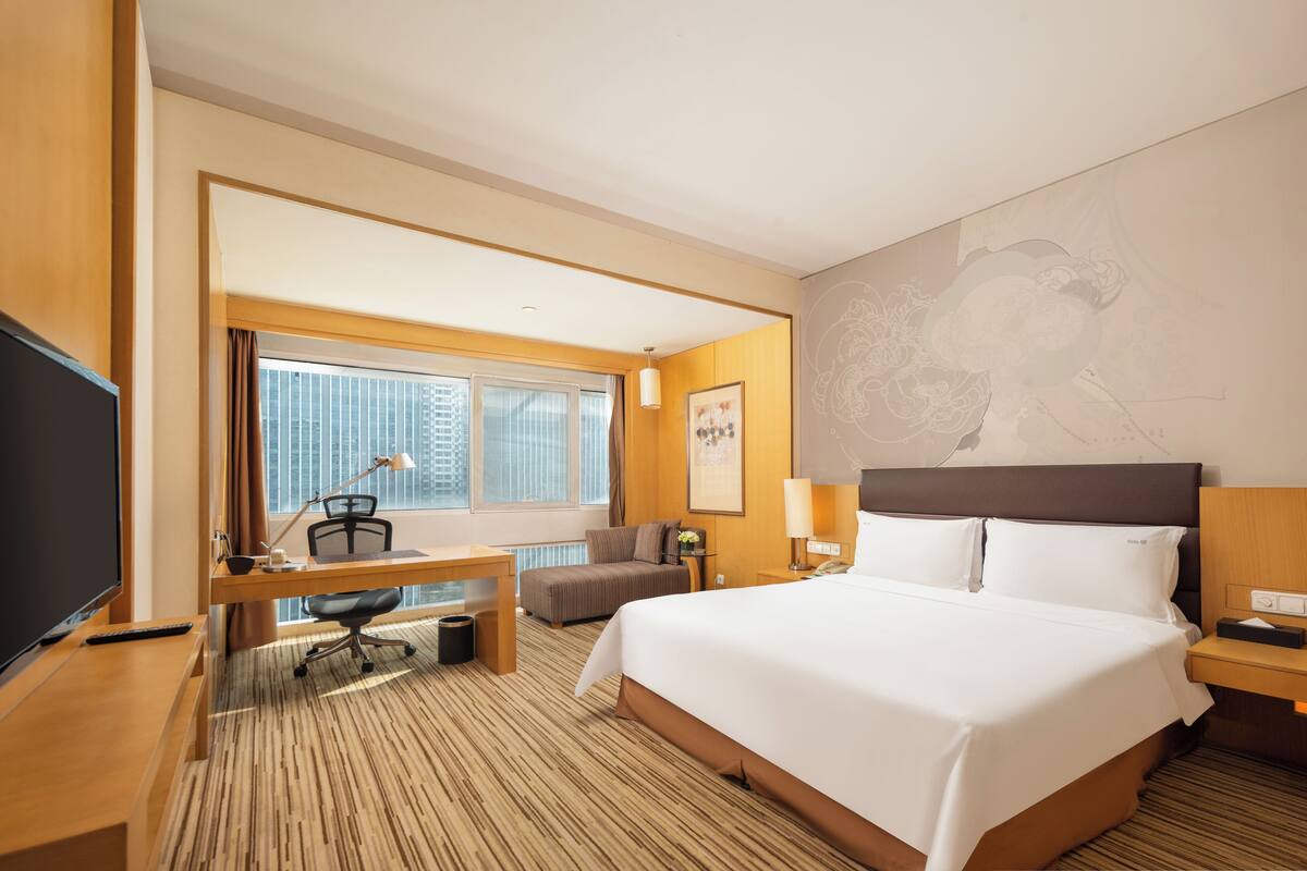 Ein Hotelzimmer mit einem großen Bett, einem Schreibtisch mit Computer, einem Sessel, einem Fernseher und einem Fenster mit Jalousien.