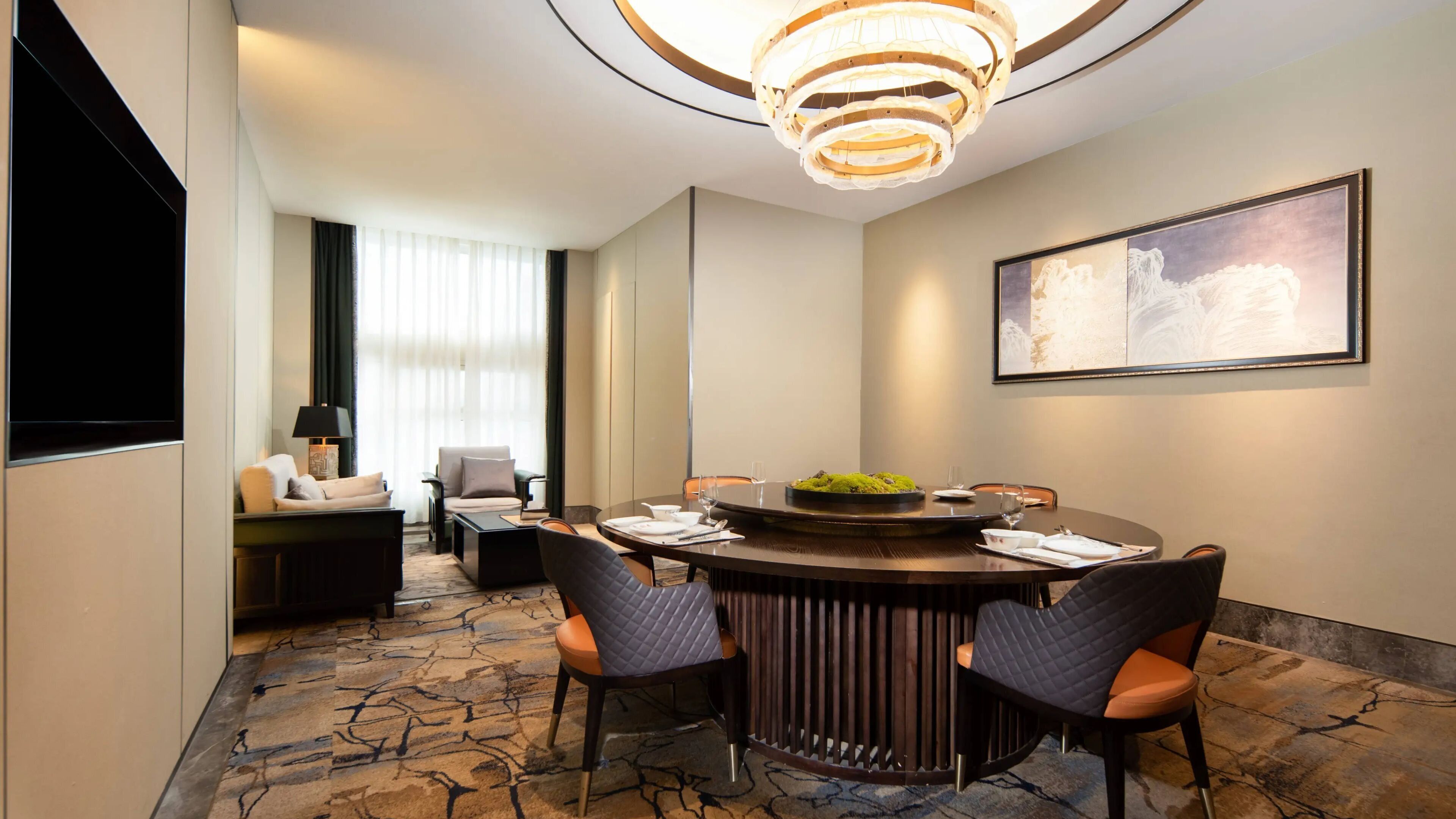 A modern hotel room with a round dining table, four chairs, a sofa, a TV, and a large painting on the wall.