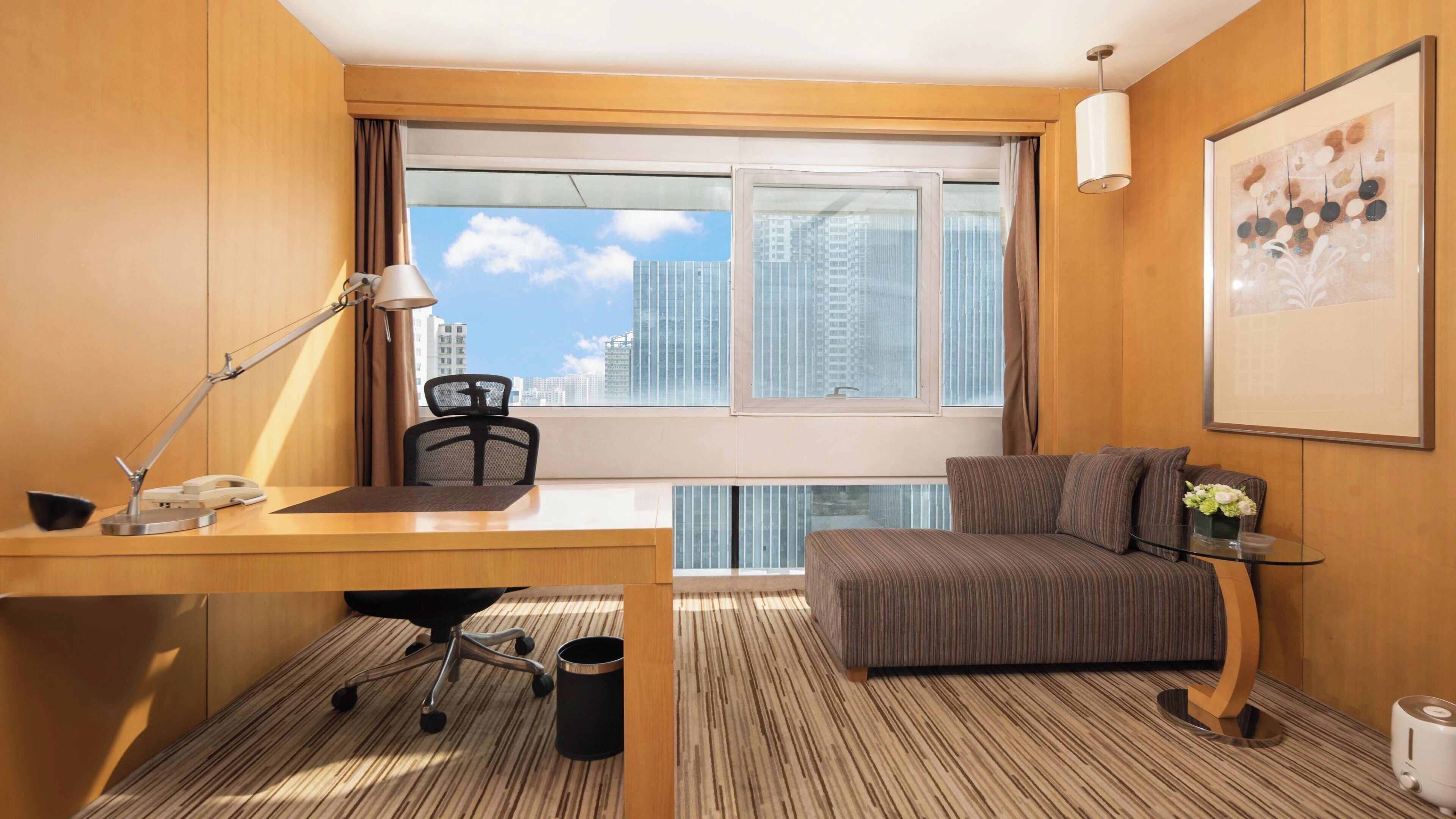 Een hotelkamer met een bureau, stoel, bank en een groot raam met uitzicht op de stad.
