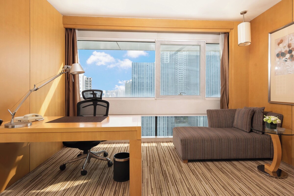 Ein Hotelzimmer mit Schreibtisch, Stuhl und einer Couch. Ein Fenster bietet Blick auf Gebäude.