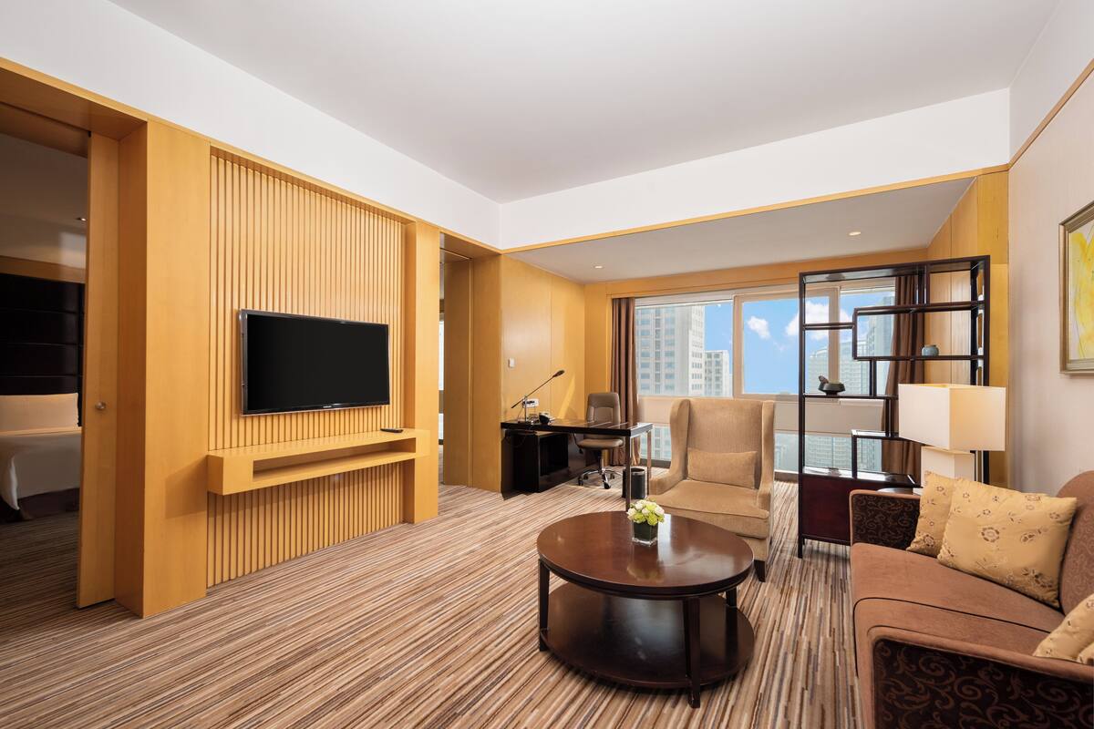Ein modernes Hotelzimmer mit großem Flachbildfernseher an einer Wand aus Holzpaneelen, einer bequemen Couch, einem runden Couchtisch und einem Schreibtisch mit Stuhl.