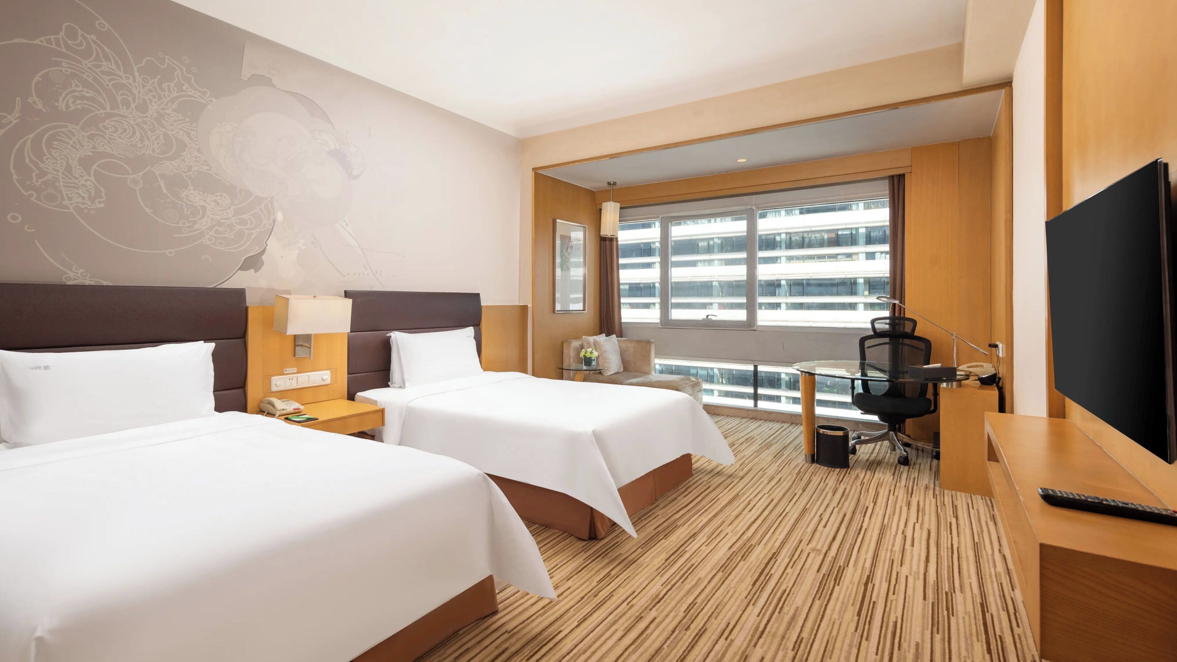 A hotel room with two beds, a desk, a TV, and a large window with a view of buildings.