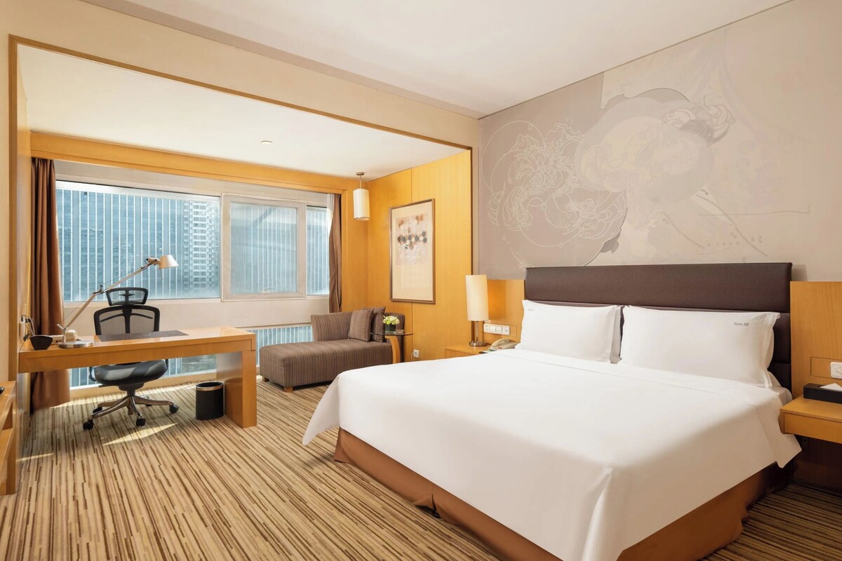 Ein Hotelzimmer mit einem großen Bett, einem Schreibtisch mit Stuhl, einem Fernseher und einem Fenster mit Vorhängen.