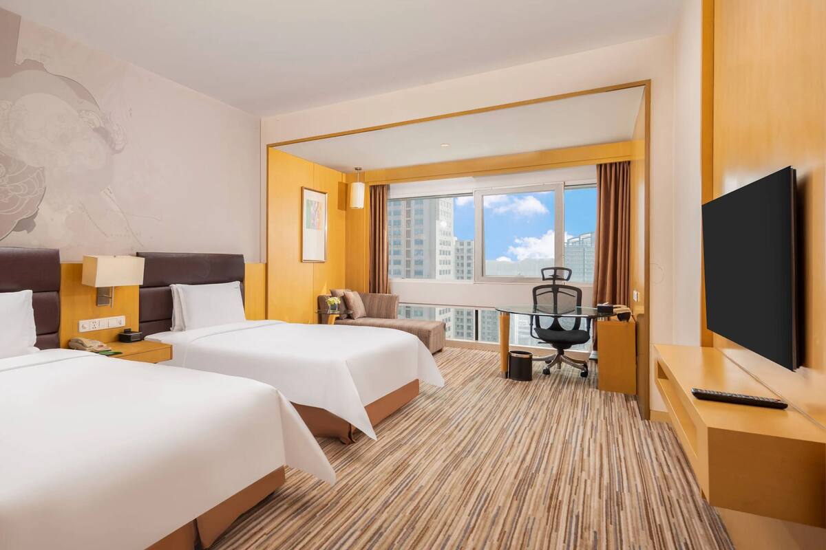 Ein Hotelzimmer mit zwei Betten, einem Schreibtisch, einem Stuhl, einem Fernseher und einem Fenster mit Blick auf die Stadt.