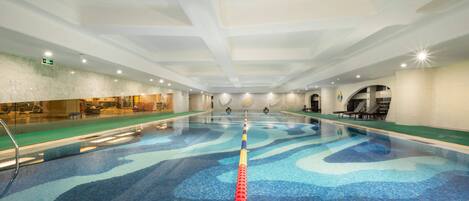 Una amplia piscina interior con un separador de carril claro, una zona de recepción con pared de vidrio y un área de descanso con sillas y una mesa.