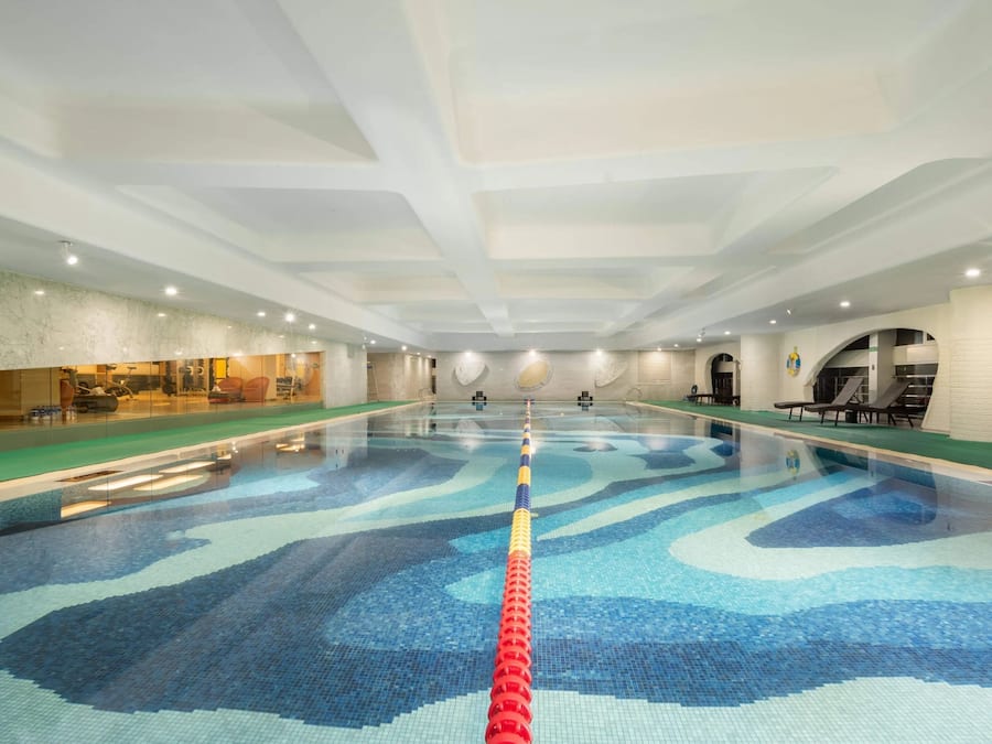 A large indoor swimming pool with a clear lane divider, a reception area with a glass wall, and a seating area with chairs and a table.