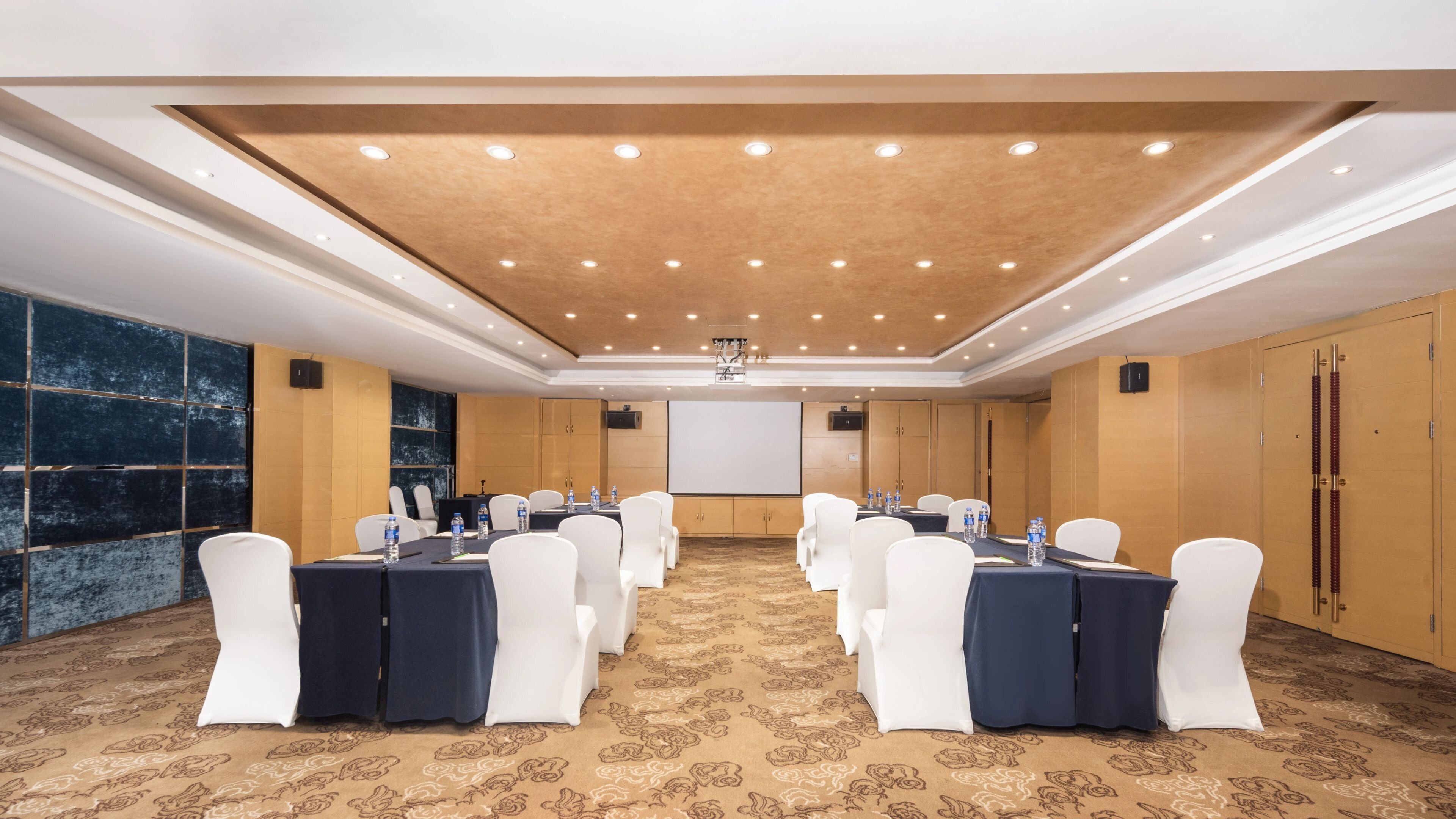A conference room with a carpeted floor, white chairs, tables with water bottles, a projector screen, and a wooden ceiling.