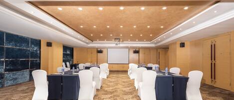 A conference room with a carpeted floor, white chairs, tables with water bottles, a projector screen, and a wooden ceiling.