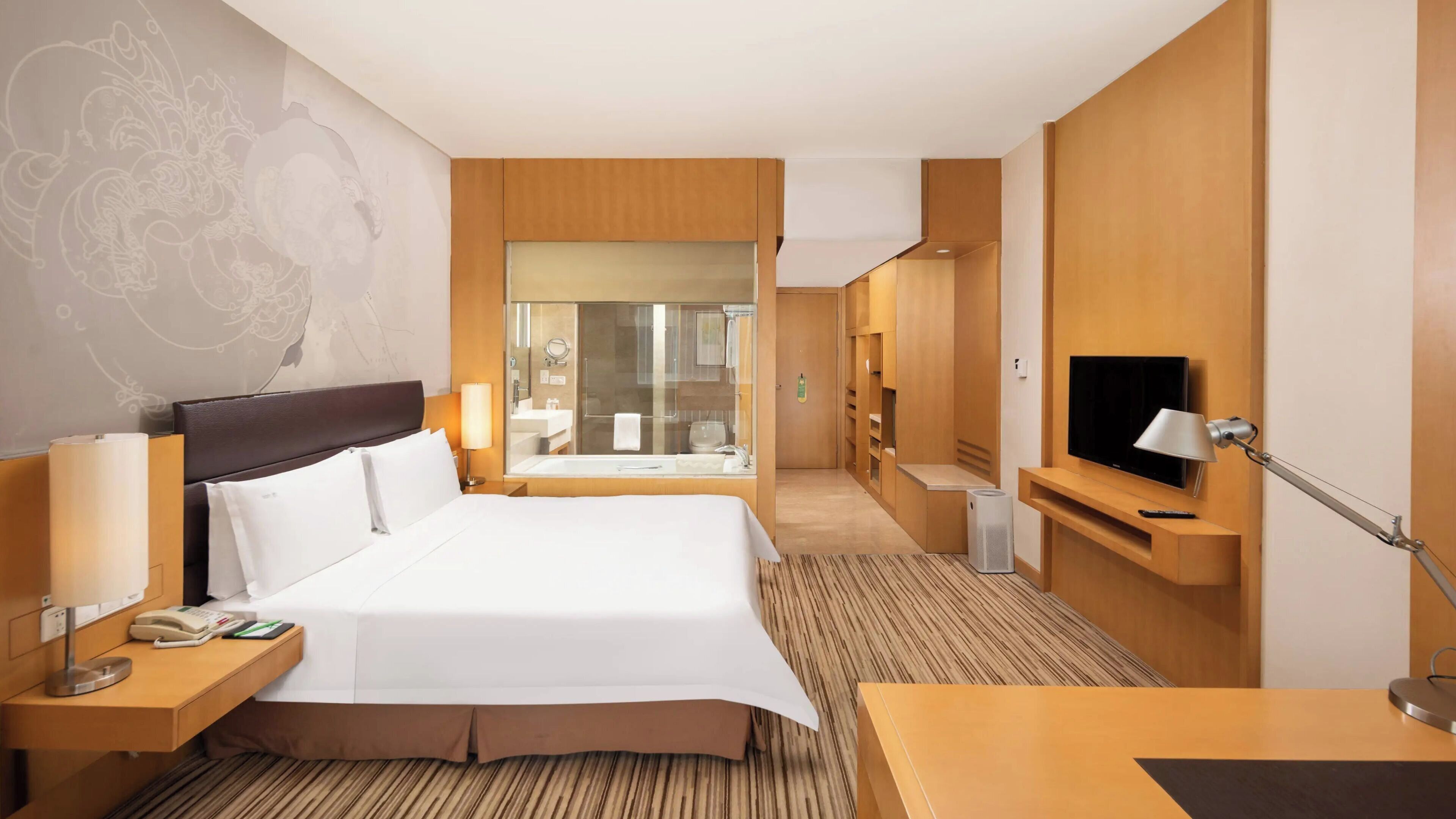 A hotel room with a large bed, bedside tables, a TV, a desk, and a bathroom visible through a glass door.