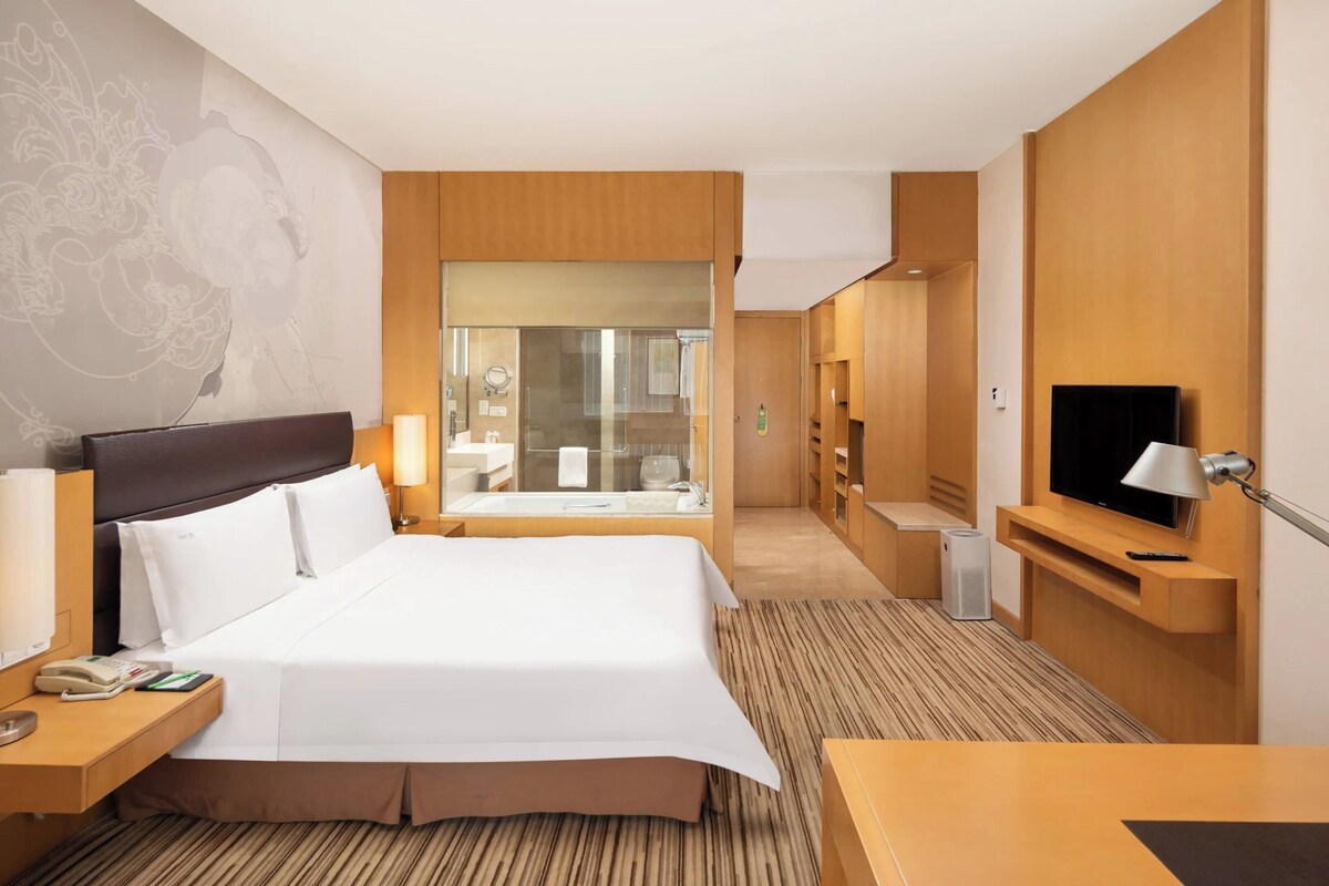Ein Hotelzimmer mit einem großen Bett, Nachttischen, einem Fernseher, einem Schreibtisch und einem durch eine Glastür einsehbaren Badezimmer.