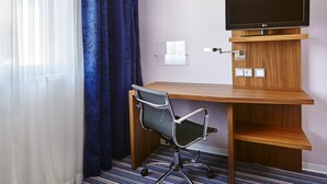 Een hotelkamer met een houten bureau, een stoel, een flatscreen televisie aan de wand en een raam met blauwe gordijnen.